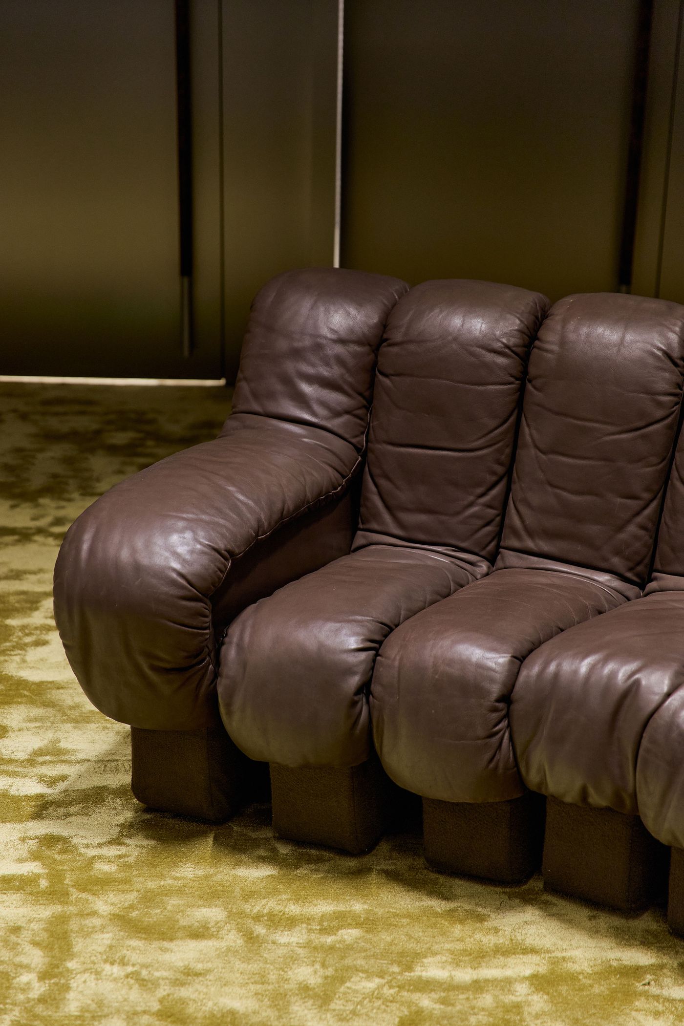 A close-up of a curved, plush brown leather sofa on a soft green carpet, highlighting its cozy, modern design.