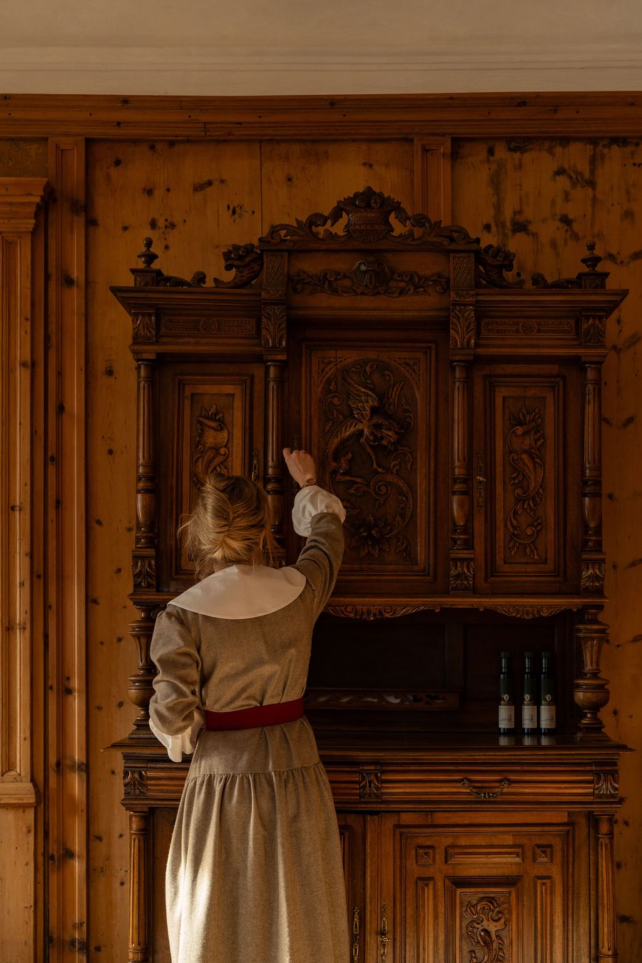 Against knotty pine panelling at Castel Badia, a server reaches towards an imposing carved walnut buffet. Its central door bears a winged griffin amid foliate scrolls, framed by turned columns, acanthus carving and a pierced crest, while three bottles of olive oil stand in the open recess below. Against knotty pine panelling at Castel Badia, a server reaches towards an imposing carved walnut buffet. Its central door bears a winged griffin amid foliate scrolls, framed by turned columns, acanthus carving and a pierced crest, while three bottles of olive oil stand in the open recess below.