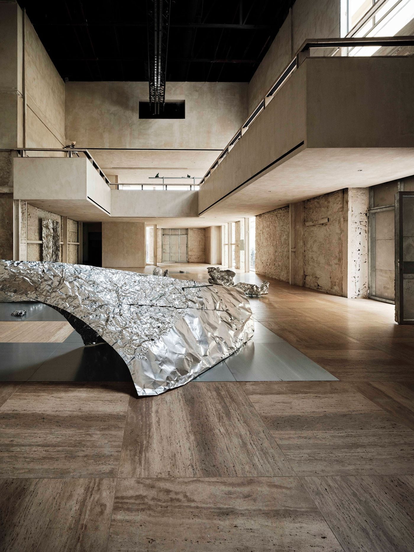 Castor Place’s main hall is animated by a large reflective installation from “Casting Water”, its crumpled metallic planes spreading across grey floor panels and the warm-toned travertine grid. Above, pale suspended galleries and the original black roof structure emphasise the dramatic height and layered spatial composition Castor Place’s main hall is animated by a large reflective installation from “Casting Water”, its crumpled metallic planes spreading across grey floor panels and the warm-toned travertine grid. Above, pale suspended galleries and the original black roof structure emphasise the dramatic height and layered spatial composition