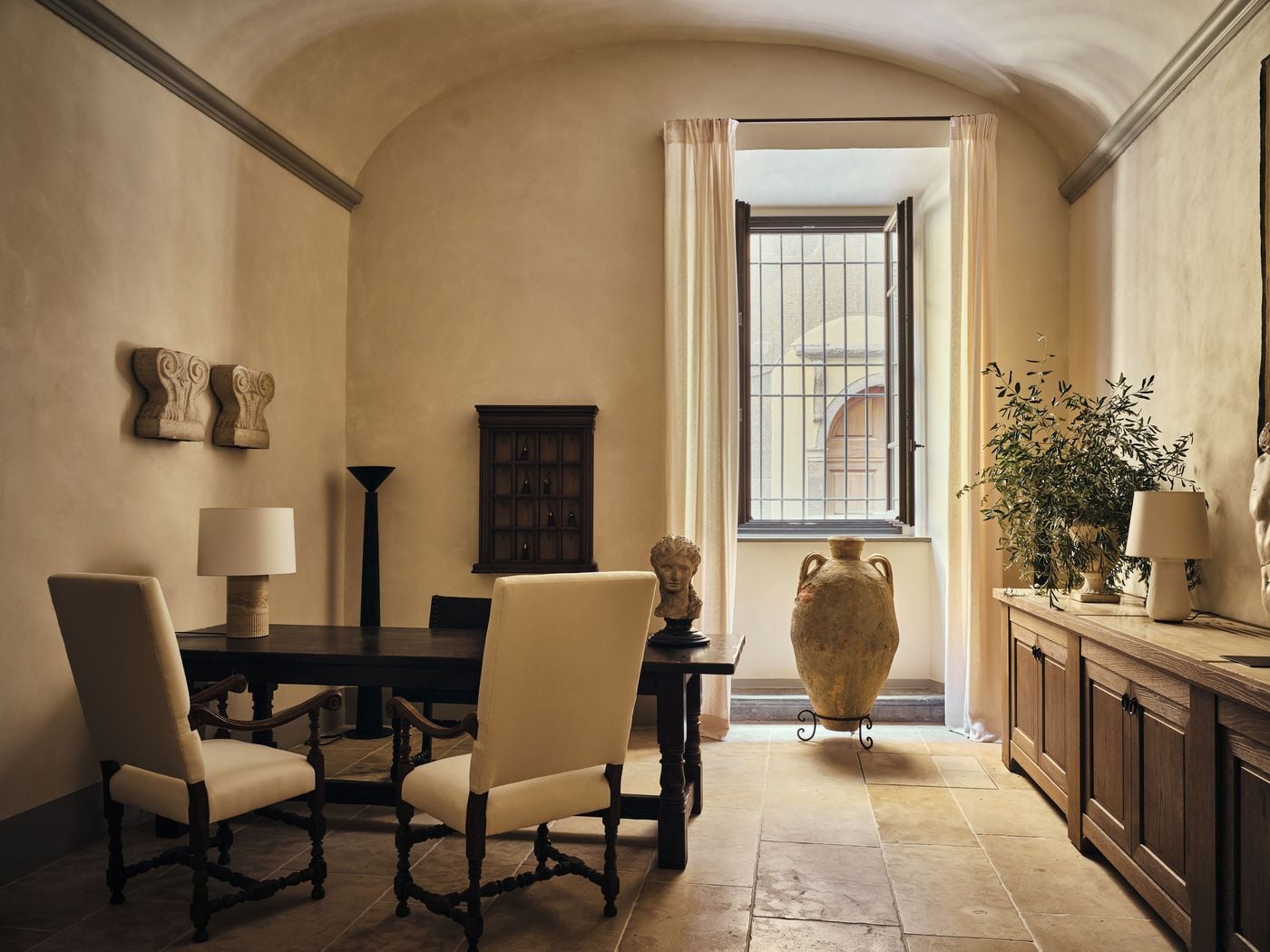 A vaulted reception room at La Réserve Firenze pairs an antique refectory table and high-backed upholstered chairs with a classical head on a stand. A wall cabinet of hanging keys, carved stone capitals, a terracotta amphora on an iron stand and a barred window onto the street complete the scene. A vaulted reception room at La Réserve Firenze pairs an antique refectory table and high-backed upholstered chairs with a classical head on a stand. A wall cabinet of hanging keys, carved stone capitals, a terracotta amphora on an iron stand and a barred window onto the street complete the scene.