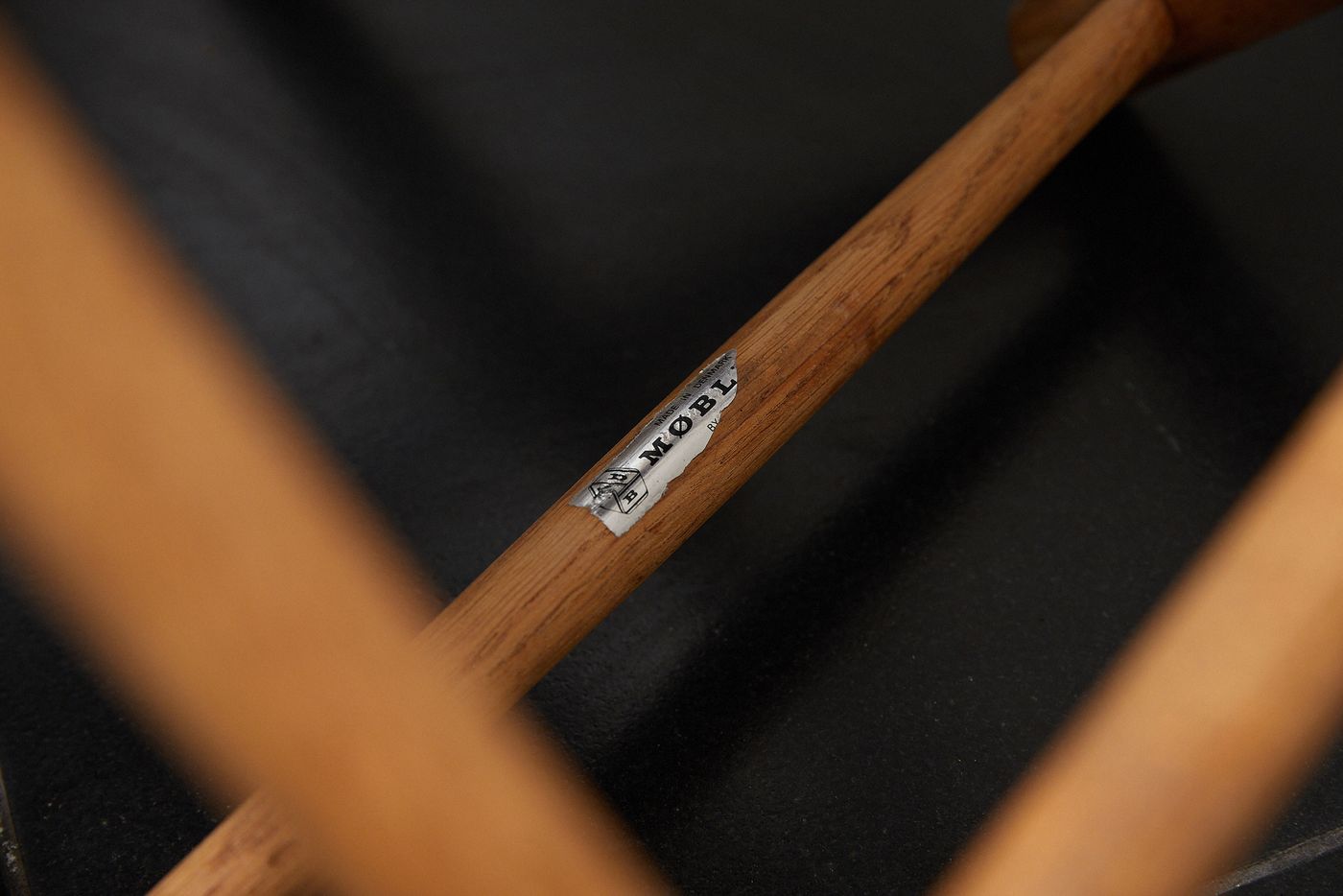 Close-up of a wooden chair leg featuring a metal label that reads "MØBL" against a dark background.