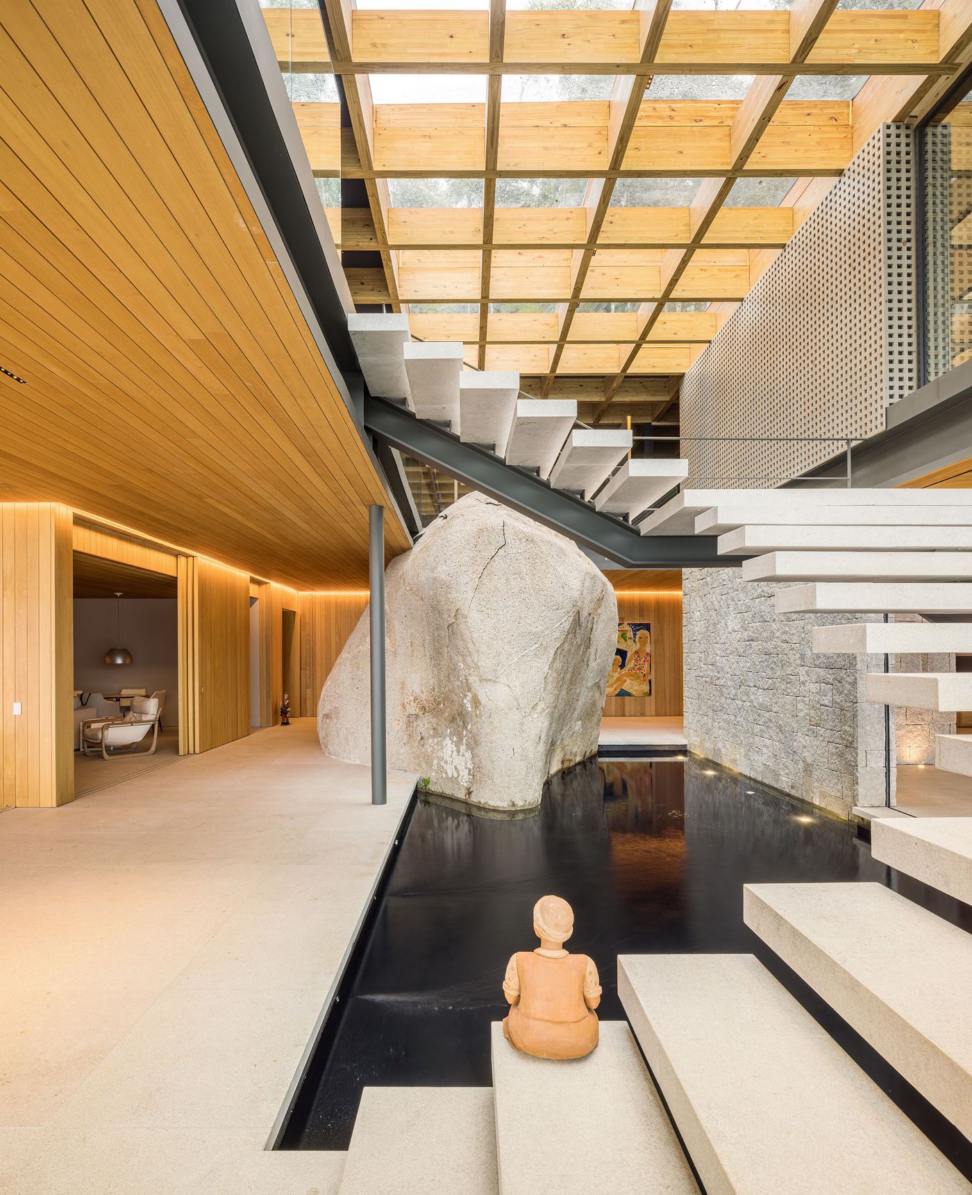 A grand interior space features an exposed timber grid ceiling and a dramatic floating staircase with a large natural rock formation. A dark, reflecting pool runs alongside, creating a serene and integrated environment, while an Eastern-inspired sculpture adds cultural depth. A grand interior space features an exposed timber grid ceiling and a dramatic floating staircase with a large natural rock formation. A dark, reflecting pool runs alongside, creating a serene and integrated environment, while an Eastern-inspired sculpture adds cultural depth.