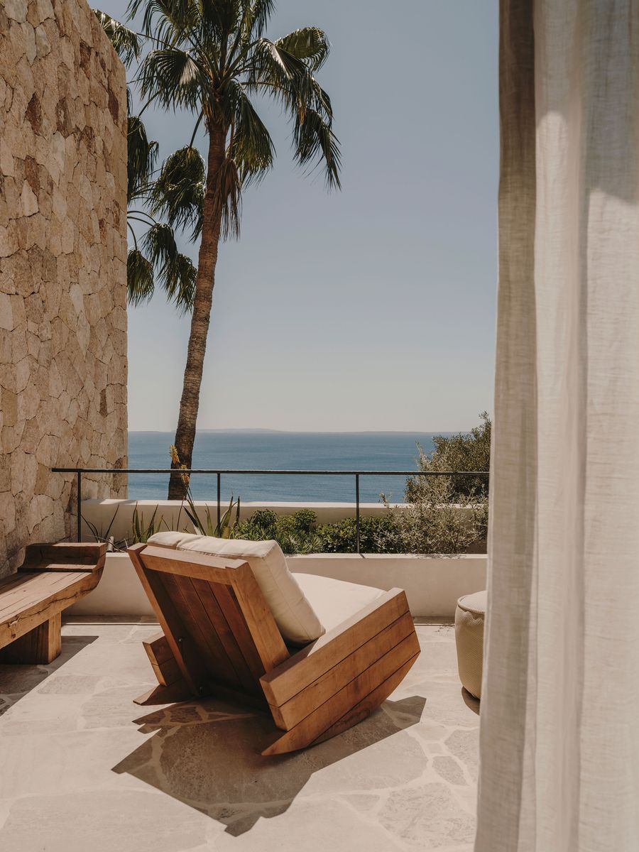 A sunlit terrace with a wooden lounge chair, overlooking a serene ocean view and a palm tree, framed by sheer white curtains. A sunlit terrace with a wooden lounge chair, overlooking a serene ocean view and a palm tree, framed by sheer white curtains.