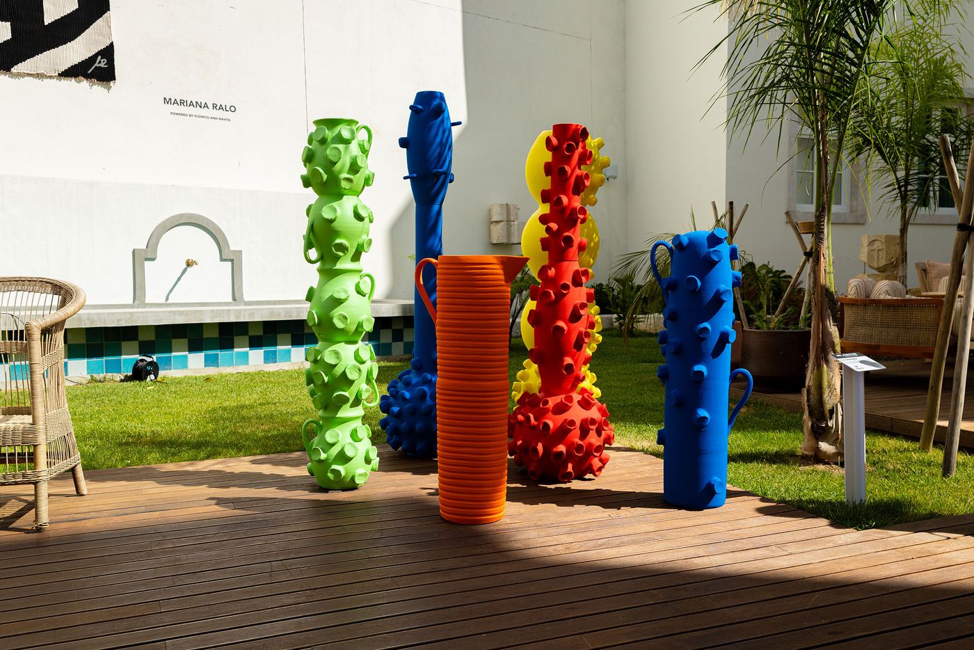 A collection of vibrant, large-scale contemporary sculptures with organic, textured surfaces by Terrakota Studio are displayed outdoors at Locke Santa Joana. Tall, colorful forms in lime green, electric blue, bright orange, and red feature raised bumps and undulating shapes, set against a green lawn and wooden decking. The modern art contrasts with traditional white architecture and palm trees. A collection of vibrant, large-scale contemporary sculptures with organic, textured surfaces by Terrakota Studio are displayed outdoors at Locke Santa Joana. Tall, colorful forms in lime green, electric blue, bright orange, and red feature raised bumps and undulating shapes, set against a green lawn and wooden decking. The modern art contrasts with traditional white architecture and palm trees.