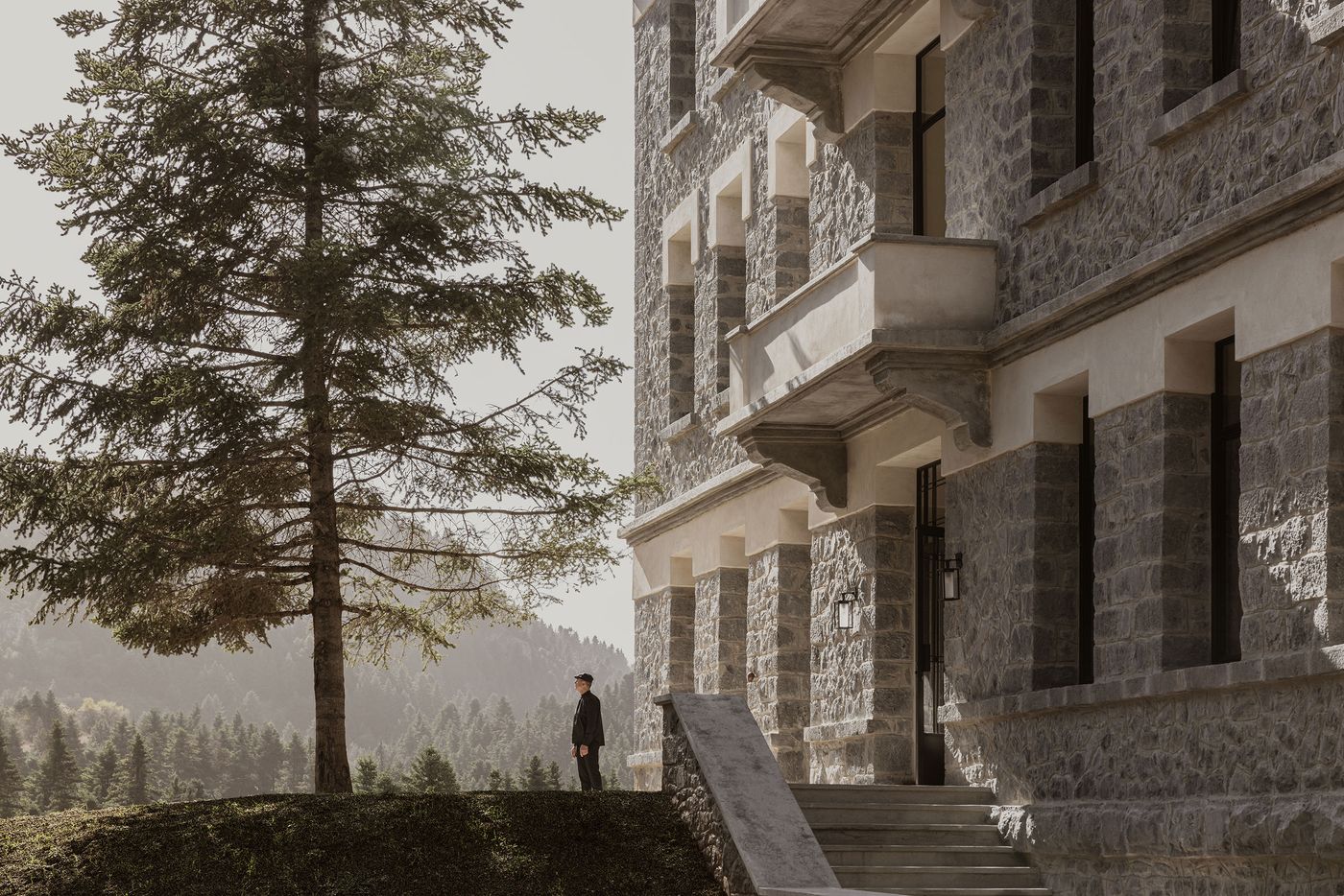 A man in a dark suit stands contemplatively on a grassy knoll, overlooking a vast, misty forest landscape, with a tall pine tree beside him. To the right, the grand stone facade of the hotel rises with its robust masonry and classic architectural details, creating a harmonious blend of human presence and natural grandeur. A man in a dark suit stands contemplatively on a grassy knoll, overlooking a vast, misty forest landscape, with a tall pine tree beside him. To the right, the grand stone facade of the hotel rises with its robust masonry and classic architectural details, creating a harmonious blend of human presence and natural grandeur.
