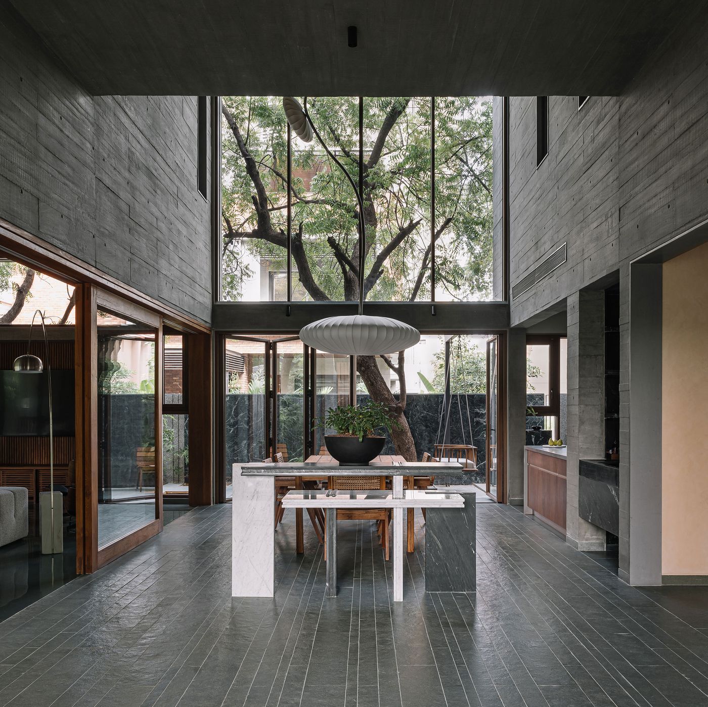 A grand double-height living space centered around a substantial tree visible through floor-to-ceiling windows. A long dining island in white and green marble adds a contemporary touch, balanced by a large, organic pendant light. Raw concrete walls and dark tiled floors create a minimalist backdrop, while glimpses of a wooden swing outside enhance the biophilic connection.