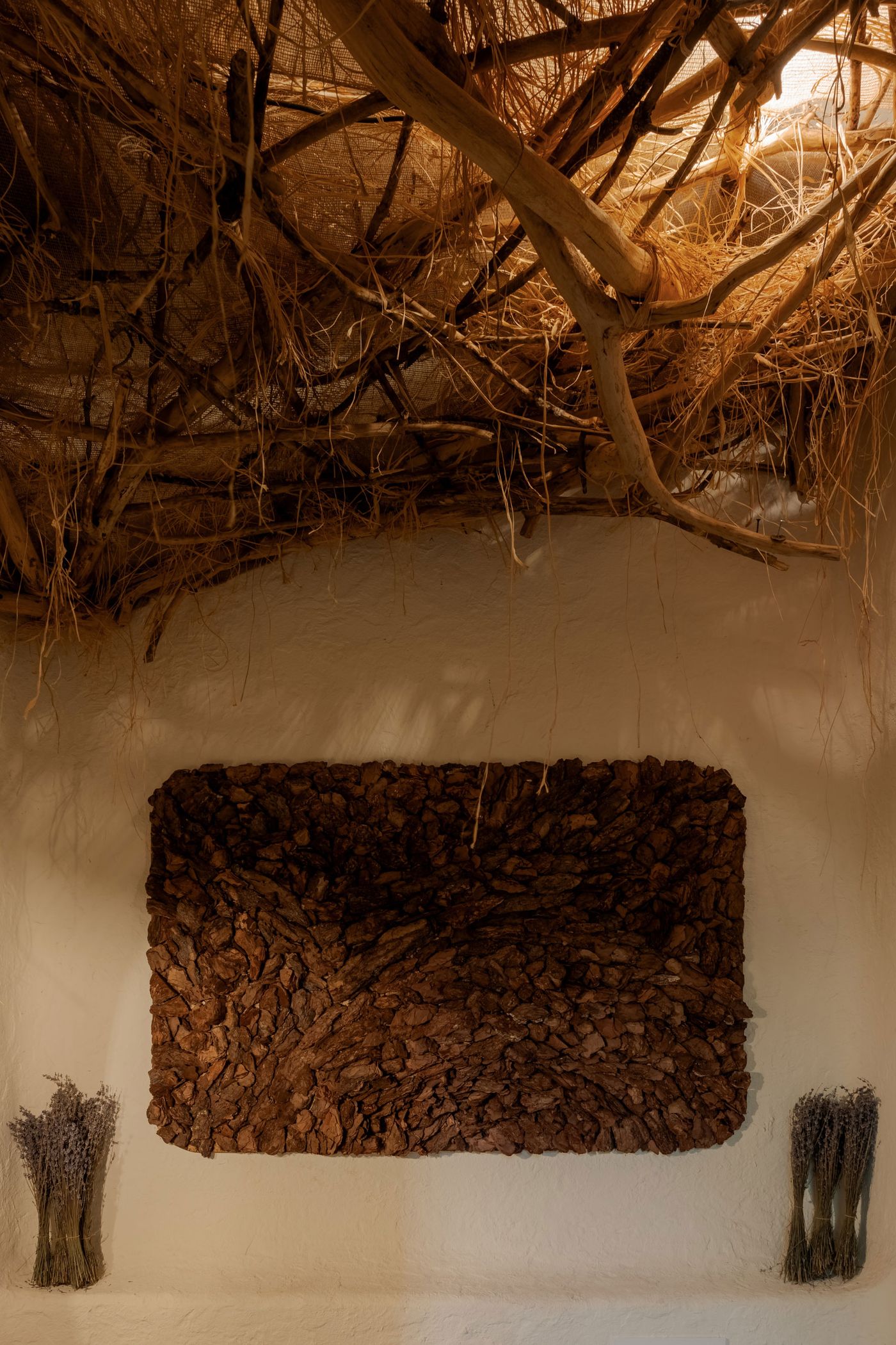 A textured brown wall sculpture resembling bark, flanked by dried lavender bouquets, under a rustic, woven natural fiber ceiling.