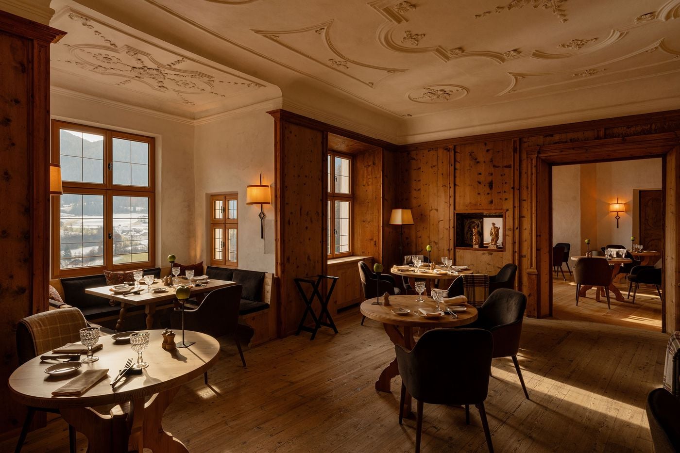 One of Castel Badia's dining rooms pairs a Baroque stucco ceiling of scrolls and floral rosettes with pine-panelled walls and deep window bays overlooking the snowy valley. Round pine tables with sculpted bases, dark upholstered armchairs, tartan throws and a niche of gilded statuettes complete the scene. One of Castel Badia's dining rooms pairs a Baroque stucco ceiling of scrolls and floral rosettes with pine-panelled walls and deep window bays overlooking the snowy valley. Round pine tables with sculpted bases, dark upholstered armchairs, tartan throws and a niche of gilded statuettes complete the scene.