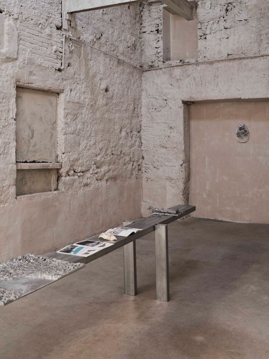 A quieter exhibition room retains Castor Place’s rough, patched masonry walls, blocked openings and exposed structural traces. A long, minimal metal display table presents books and material studies, its precise industrial geometry providing a deliberate counterpoint to the irregular, time-worn fabric surrounding it. A quieter exhibition room retains Castor Place’s rough, patched masonry walls, blocked openings and exposed structural traces. A long, minimal metal display table presents books and material studies, its precise industrial geometry providing a deliberate counterpoint to the irregular, time-worn fabric surrounding it.