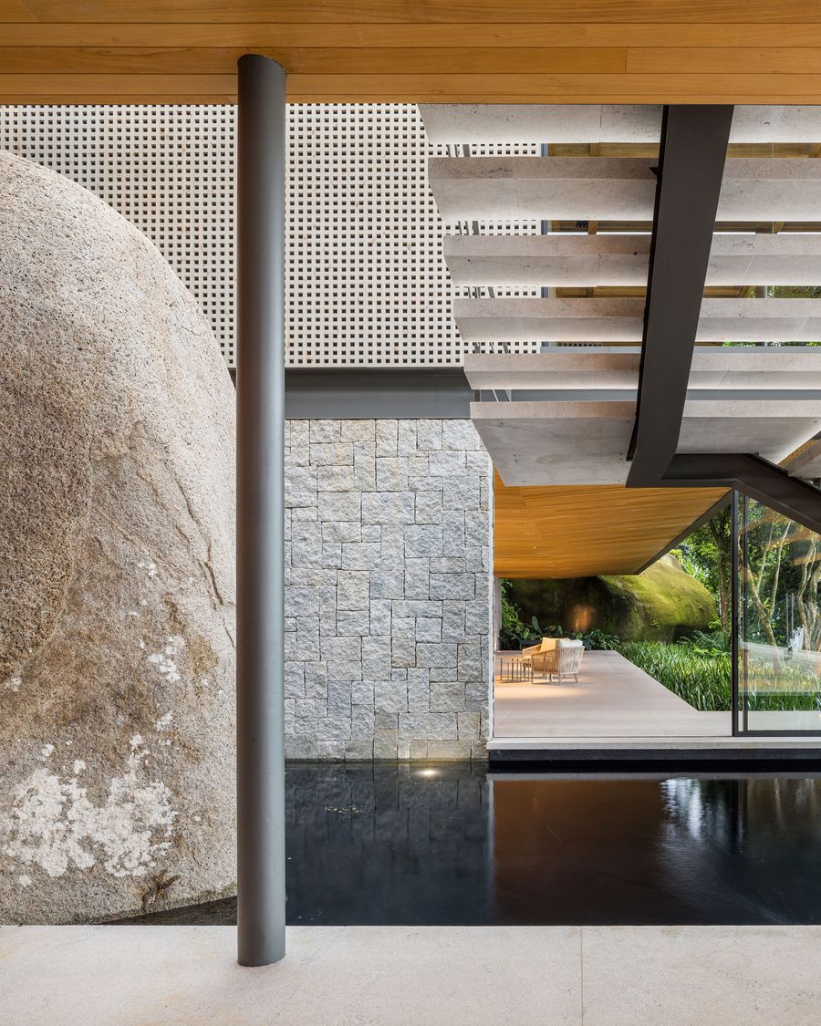 A massive, textured boulder and a stacked stone wall anchor a minimalist interior space, featuring a dark reflective pool. A striking concrete staircase with dark metal framing ascends, while perforated metal panels and warm wooden ceilings add texture and light, creating a dramatic interplay of natural and architectural elements. A massive, textured boulder and a stacked stone wall anchor a minimalist interior space, featuring a dark reflective pool. A striking concrete staircase with dark metal framing ascends, while perforated metal panels and warm wooden ceilings add texture and light, creating a dramatic interplay of natural and architectural elements.