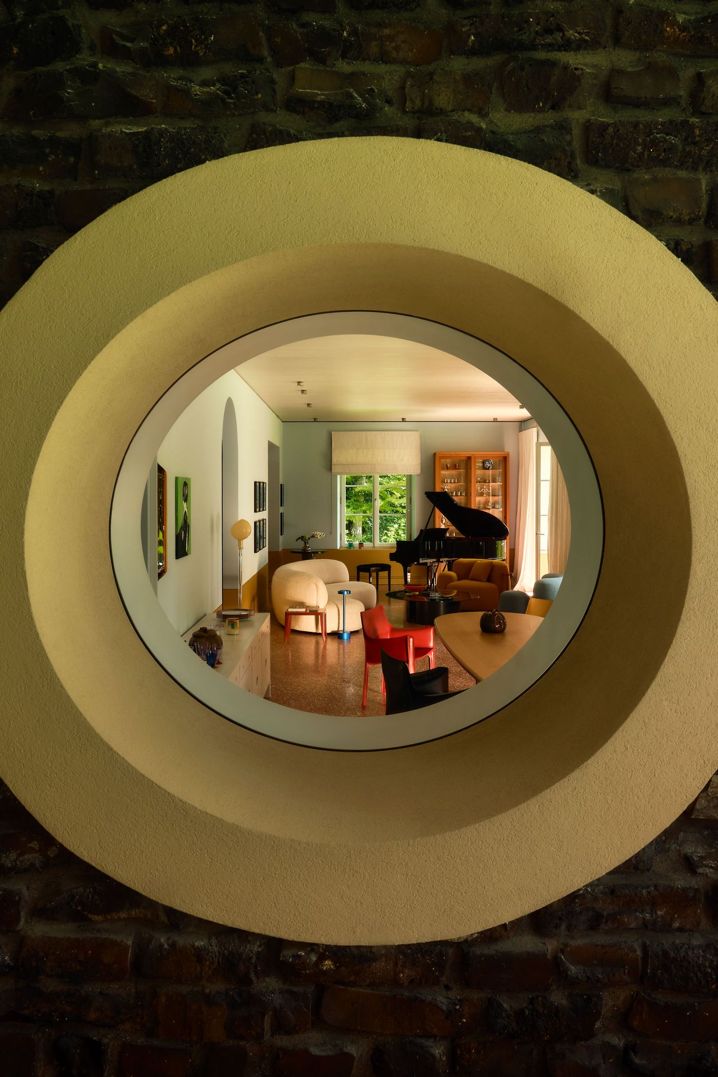 Interior view through a round window framed by textured stone, showcasing a stylish living room with colorful furniture and natural light. Interior view through a round window framed by textured stone, showcasing a stylish living room with colorful furniture and natural light.