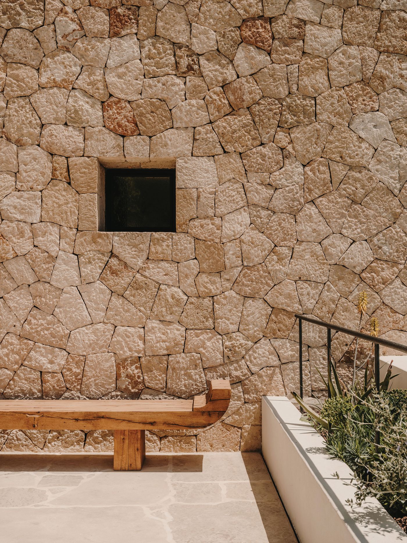 A rustic wooden bench rests against a textured stone wall, featuring a small black window and surrounding greenery. A rustic wooden bench rests against a textured stone wall, featuring a small black window and surrounding greenery.