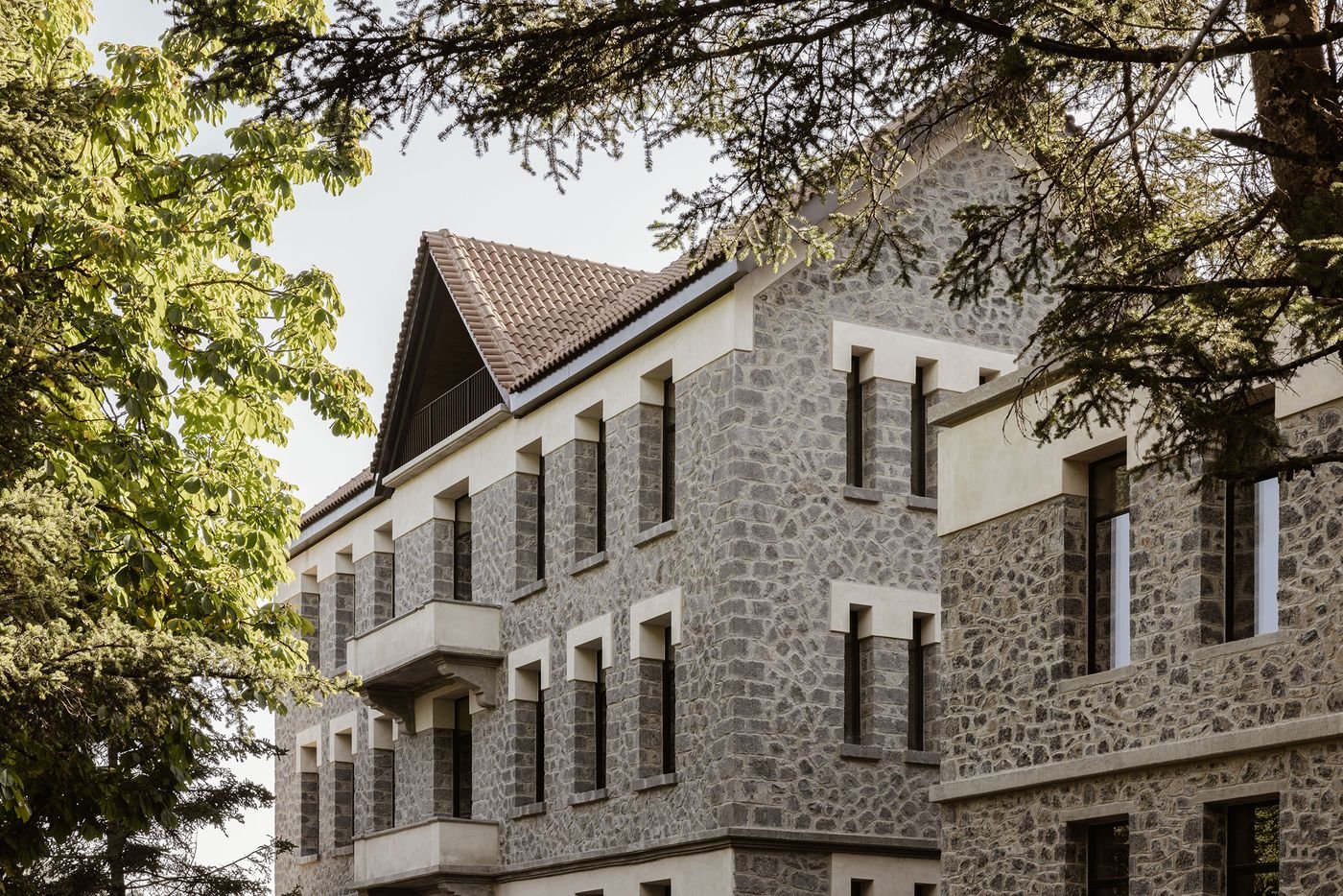 A grand hotel building constructed with textured grey stone masonry and contrasting lighter window frames, set against a bright sky. The prominent peaked roof and classical architectural details are partially framed by the lush green branches of surrounding trees, highlighting its blend of robust construction and natural integration. A grand hotel building constructed with textured grey stone masonry and contrasting lighter window frames, set against a bright sky. The prominent peaked roof and classical architectural details are partially framed by the lush green branches of surrounding trees, highlighting its blend of robust construction and natural integration.