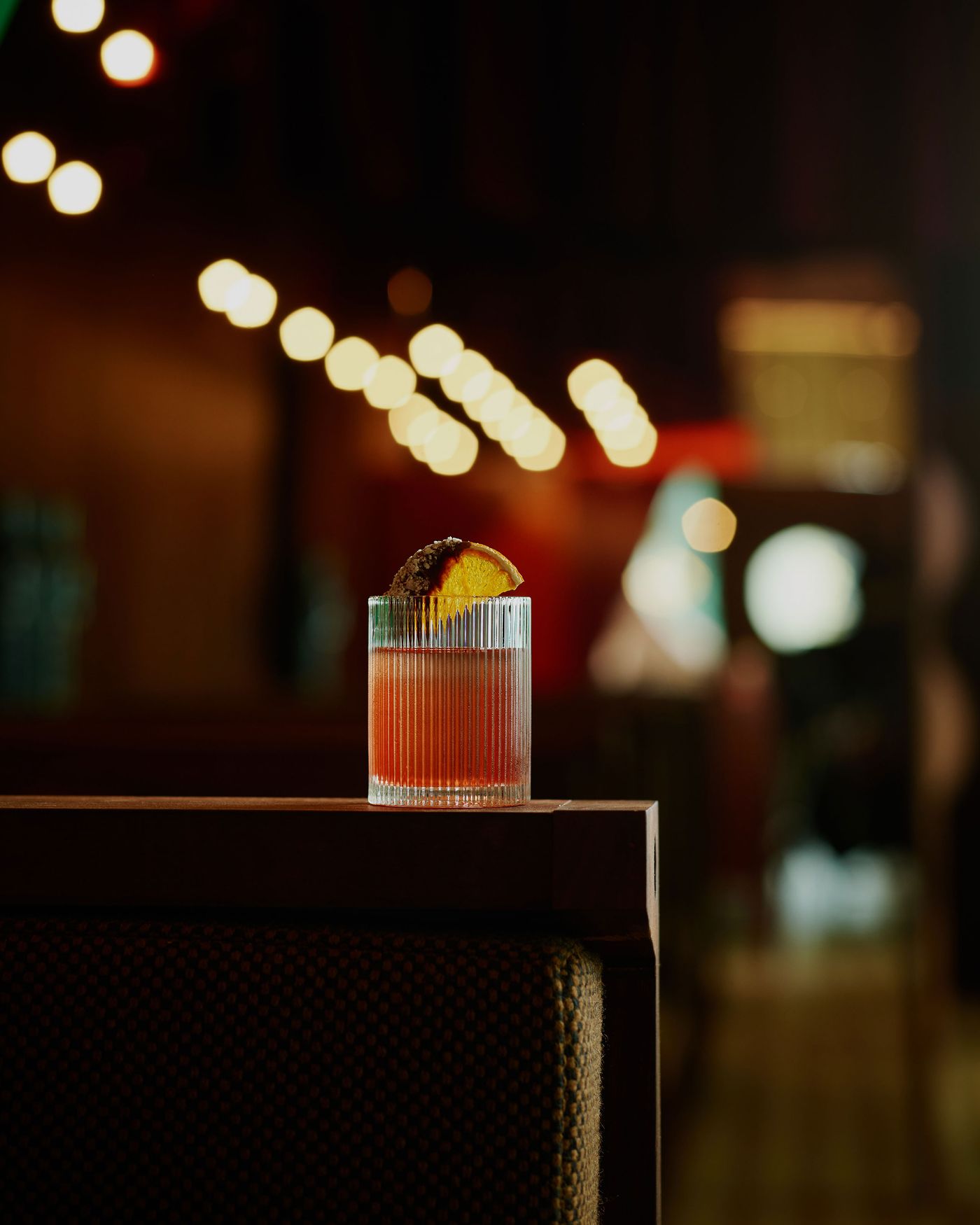 A beautifully crafted cocktail sits on a wooden table, garnished with a slice of orange, with soft bokeh lights in the background. A beautifully crafted cocktail sits on a wooden table, garnished with a slice of orange, with soft bokeh lights in the background.
