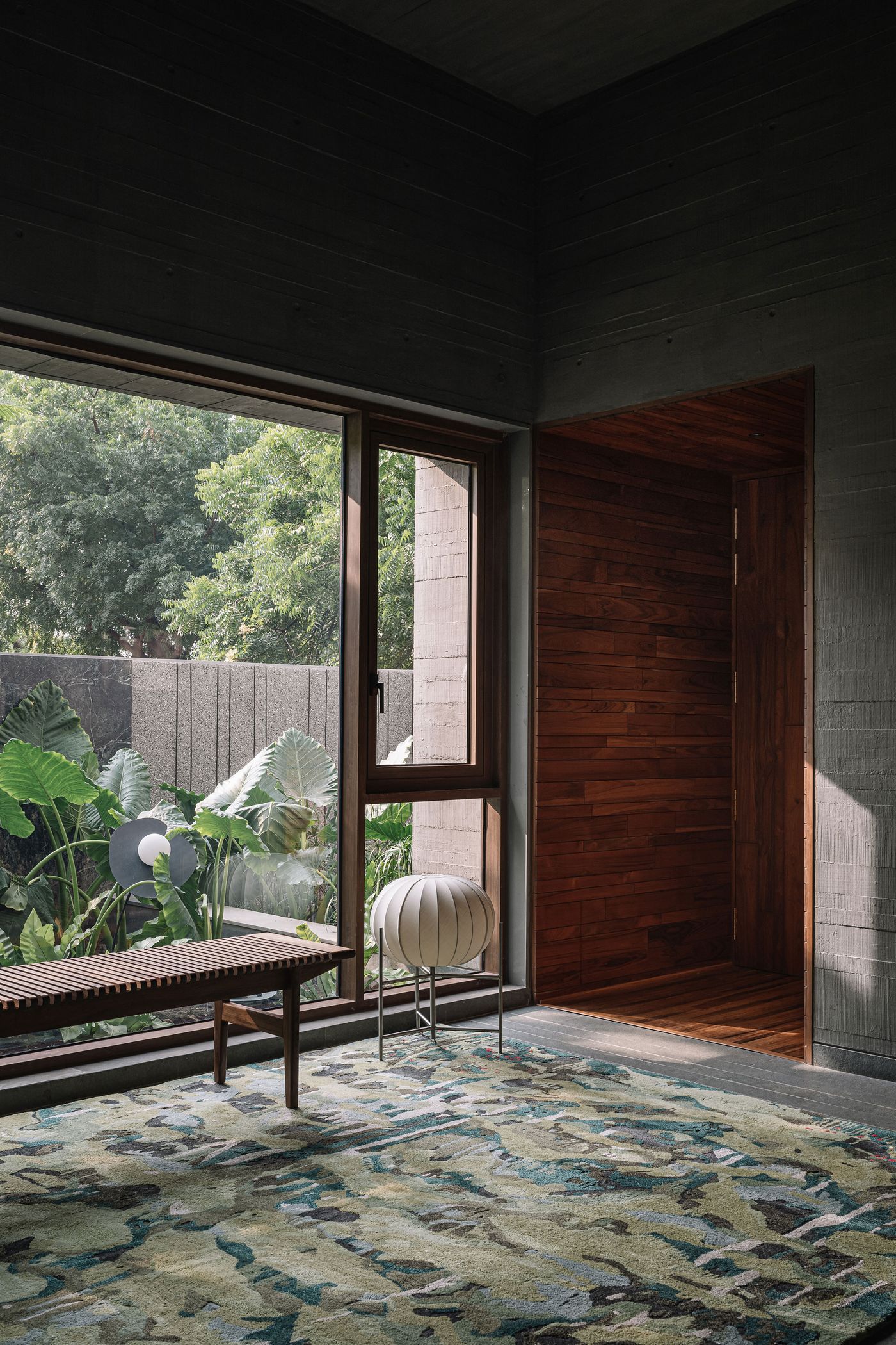 A cozy reading nook featuring a dark wooden slatted bench and a sculptural white fabric stool. The space is softened by a large, abstract rug with organic patterns in earthy green and grey tones. Expansive windows frame a view of lush outdoor plants and a textured concrete wall, creating a harmonious blend of indoor comfort and biophilic design.