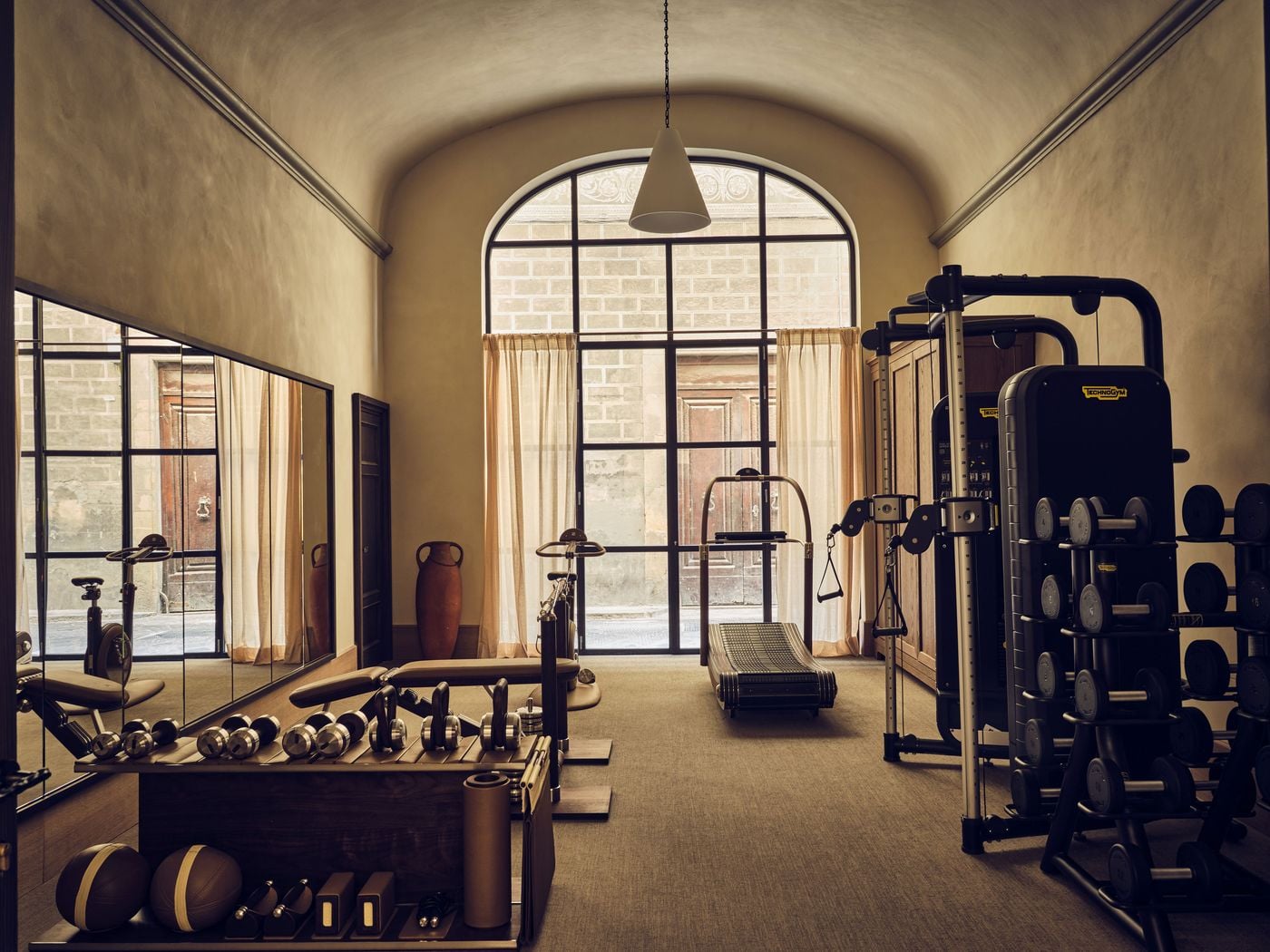 The gym at La Réserve Firenze occupies a barrel-vaulted room lit by a tall arched steel-framed window onto the street. Technogym equipment, a curved manual treadmill, timber weight racks and a mirrored wall share the space with a terracotta amphora and linen curtains, softening the functional brief. The gym at La Réserve Firenze occupies a barrel-vaulted room lit by a tall arched steel-framed window onto the street. Technogym equipment, a curved manual treadmill, timber weight racks and a mirrored wall share the space with a terracotta amphora and linen curtains, softening the functional brief.