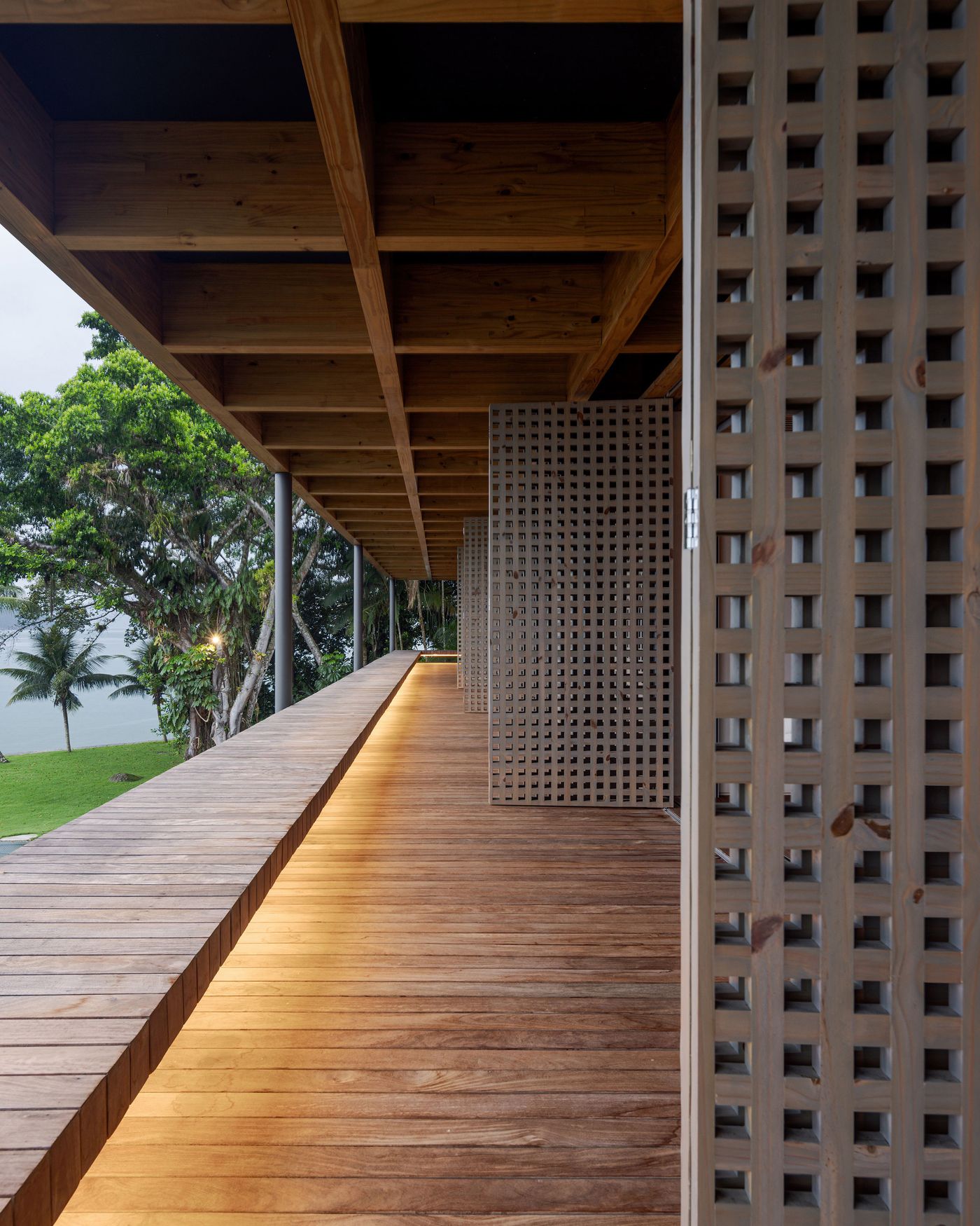 A long, elevated wooden walkway with integrated lighting extends alongside a series of perforated screens, offering a journey through the residence. The exposed timber ceiling above and distant tropical views emphasize the blend of natural materials and thoughtful design, creating a seamless connection to the outdoors.