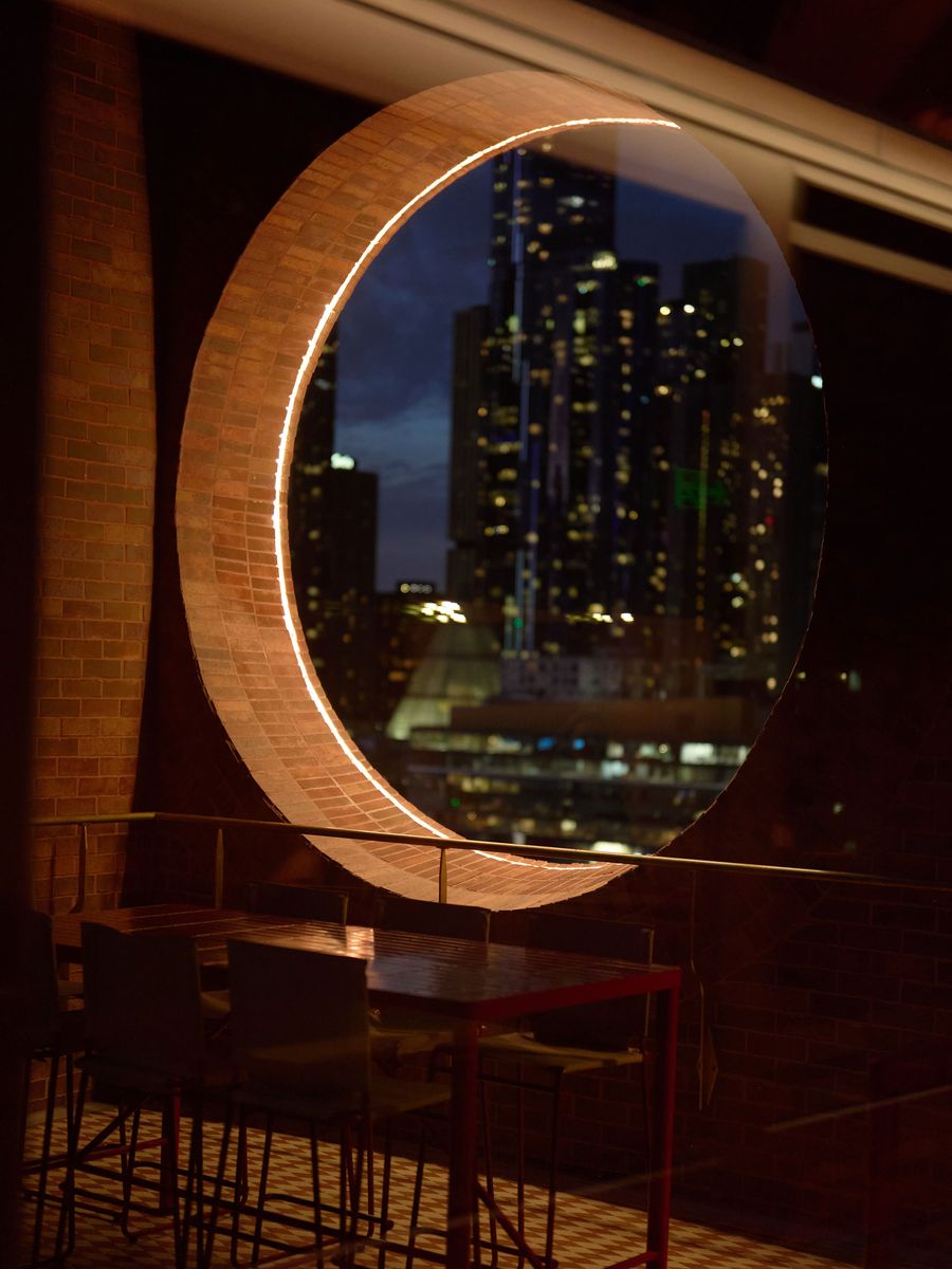 A circular window framed by brick reveals a city skyline at night, with illuminated buildings and a modern table and chairs in the foreground. A circular window framed by brick reveals a city skyline at night, with illuminated buildings and a modern table and chairs in the foreground.