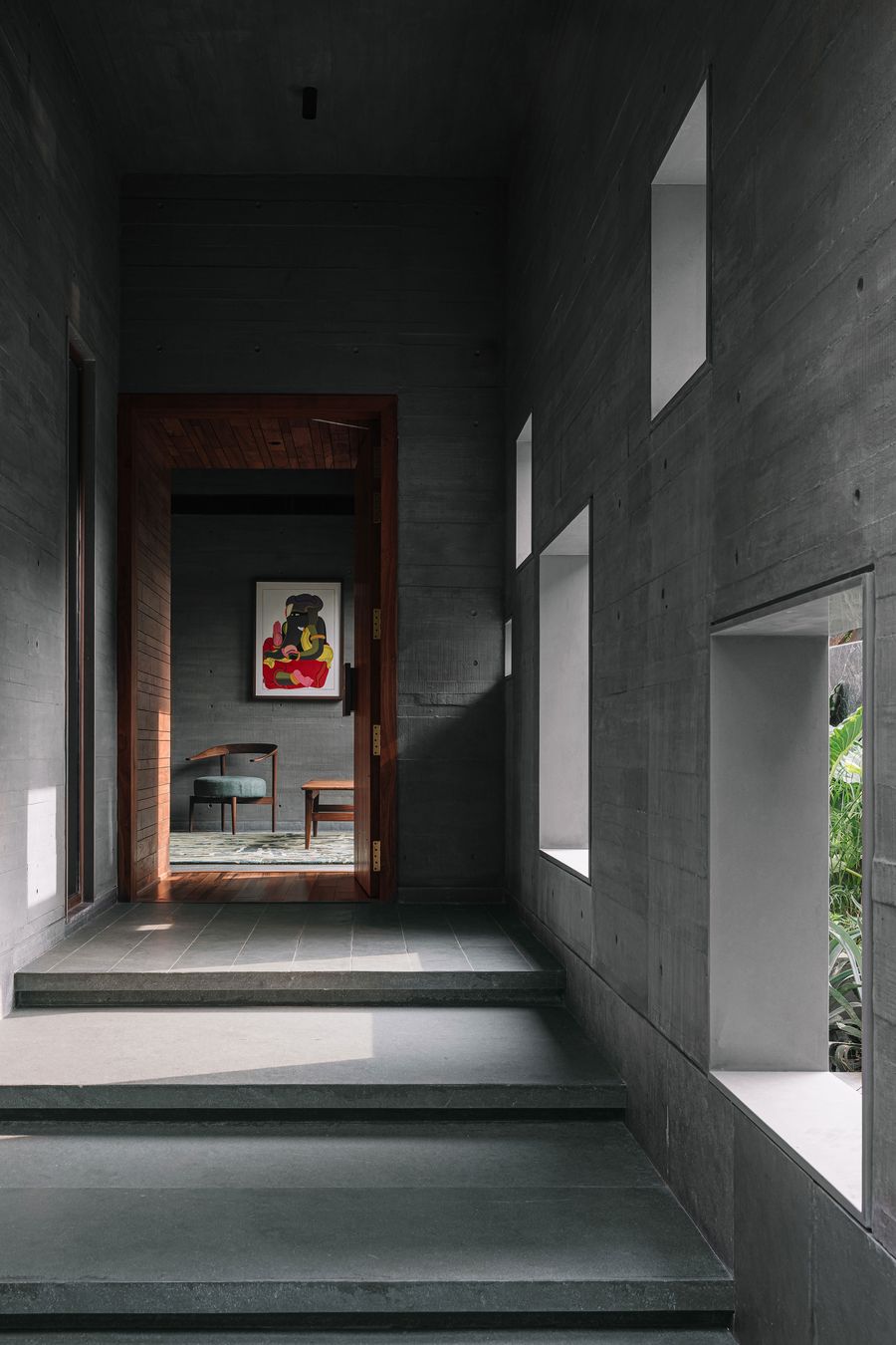 A dramatic hallway with dark concrete walls and a polished floor, leading towards a brightly lit doorway that reveals a vibrant abstract painting. A series of rectangular cutouts in the wall provide glimpses of exterior greenery and natural light, creating a dynamic interplay of light, shadow, and architectural forms.