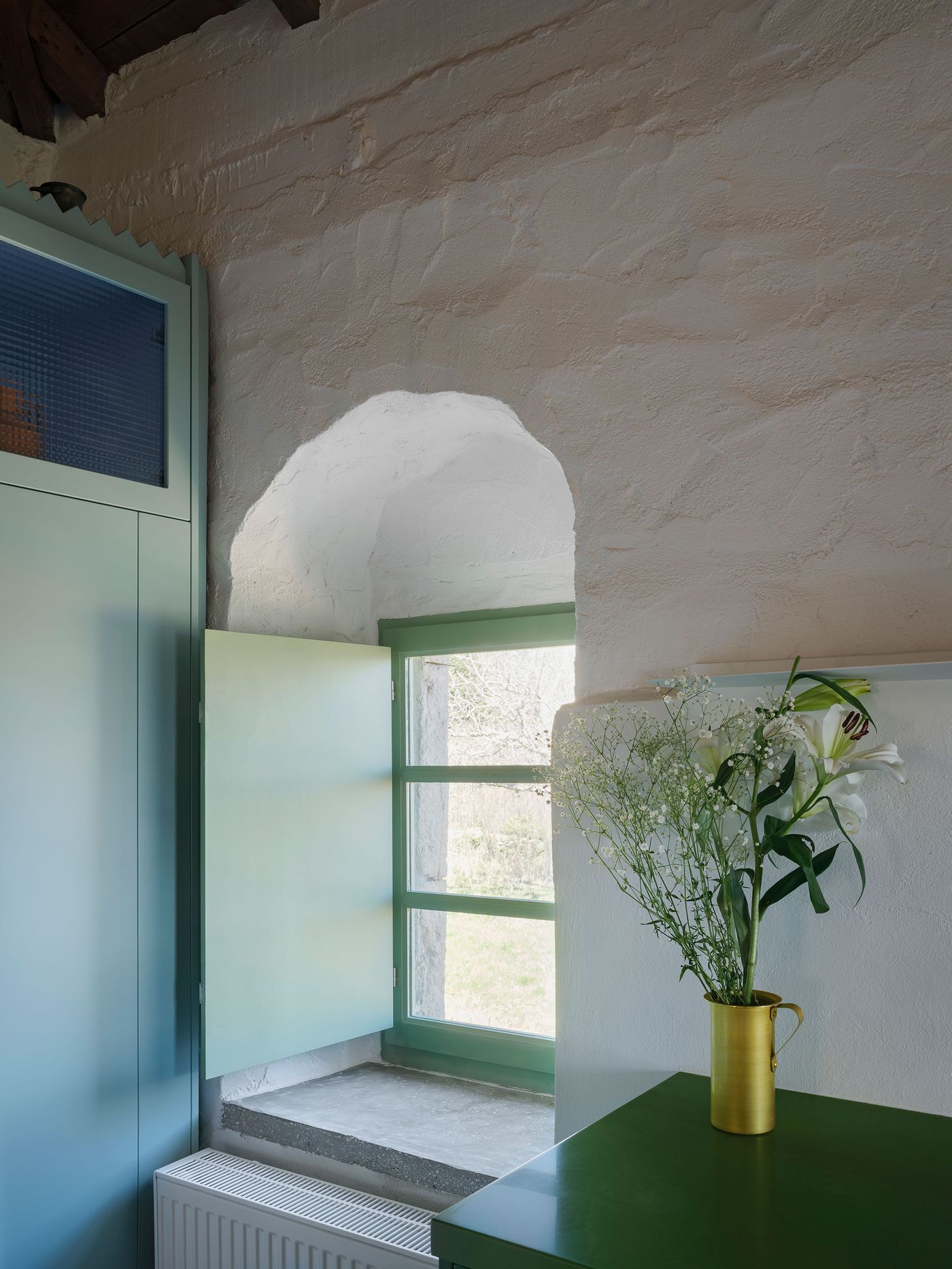 Deep arched window reveal frames the Mediterranean landscape, where sage-green contemporary elements complement the traditional whitewashed stone construction. The thickness of the walls demonstrates authentic vernacular building techniques while modern interventions provide necessary functionality without compromising the building's inherent architectural character.