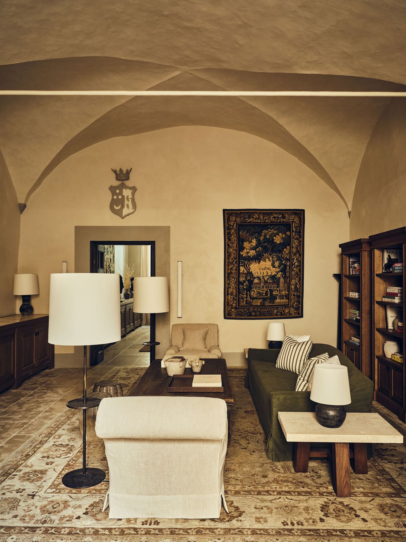 Cross vaults spring over a ground-floor lounge at La Réserve Firenze, where a painted coat of arms crowned in grey and an antique verdure tapestry anchor the walls. A moss-green slipcovered sofa, linen armchairs, oak bookcases, a travertine-topped side table and a faded patterned rug create an atmosphere of lived-in scholarship. Cross vaults spring over a ground-floor lounge at La Réserve Firenze, where a painted coat of arms crowned in grey and an antique verdure tapestry anchor the walls. A moss-green slipcovered sofa, linen armchairs, oak bookcases, a travertine-topped side table and a faded patterned rug create an atmosphere of lived-in scholarship.