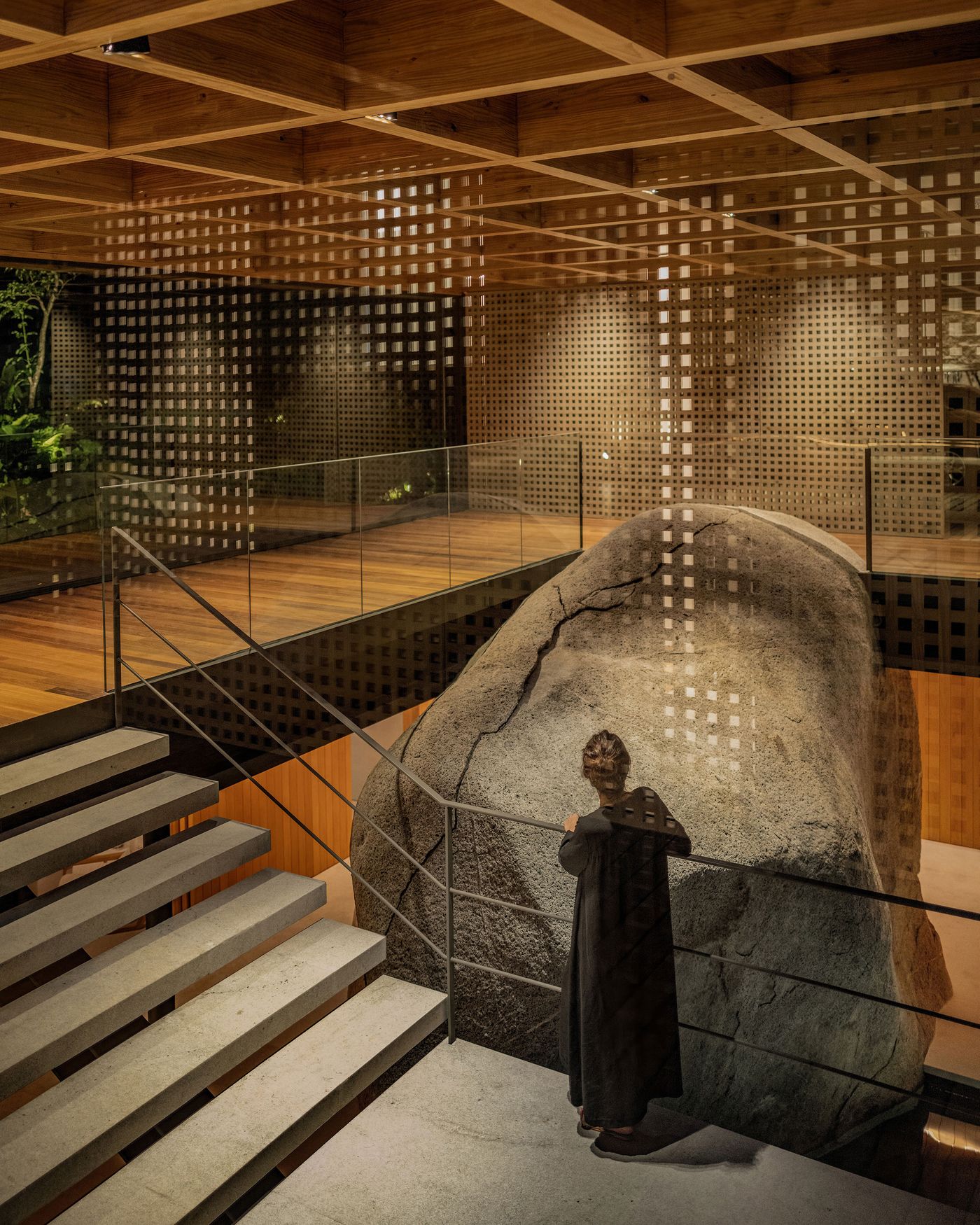 A woman in a dark dress stands on a concrete staircase, looking towards a monumental natural rock formation integrated into the interior. The space is defined by warm wooden floors, intricate timber ceilings, and perforated metal screens, all illuminated by soft ambient lighting, creating a tranquil fusion of natural and refined elements.