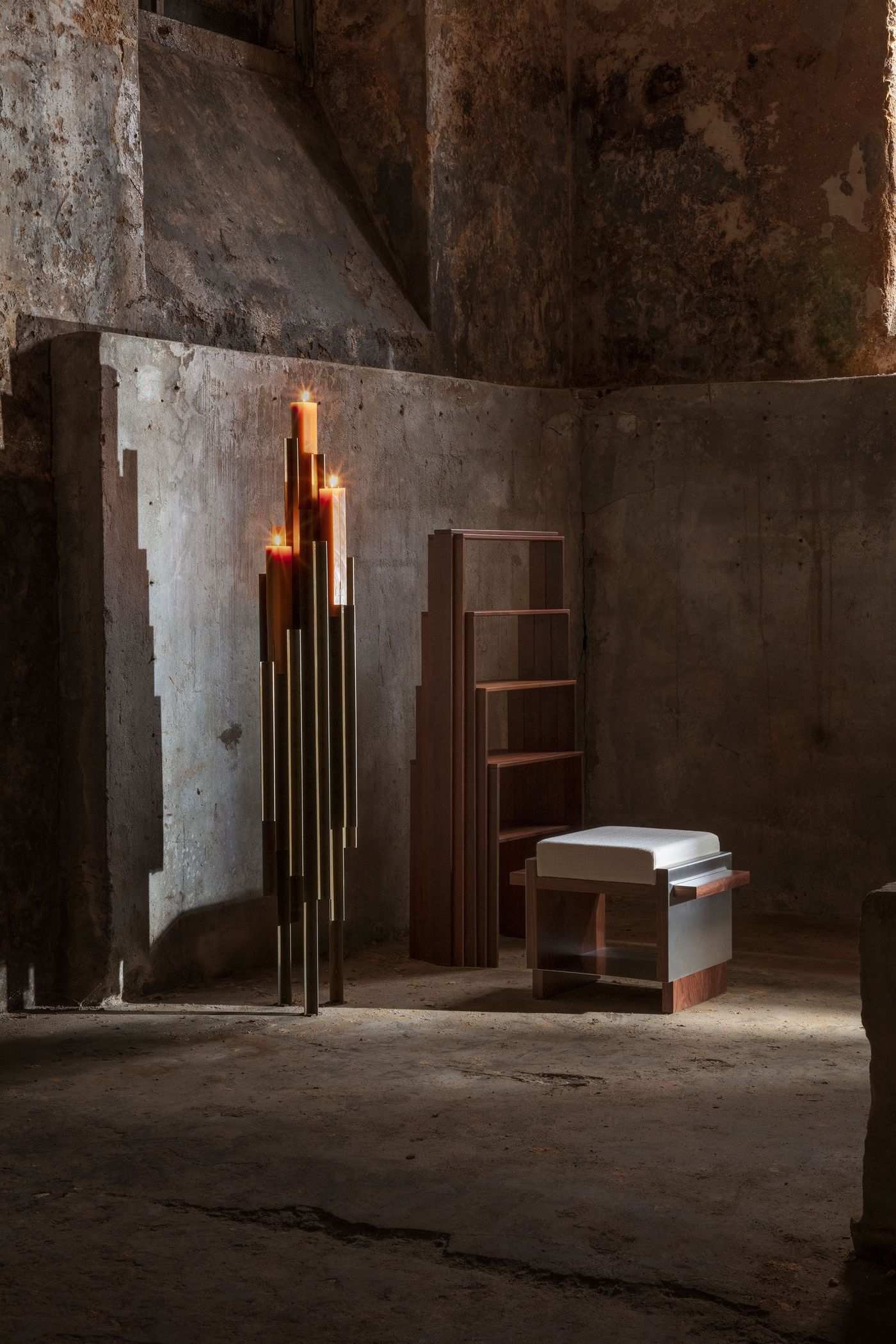 A captivating display from the "Perspective and Matter" exhibition by Luso Collective set within a raw, industrial space. A tall, sculptural floor lamp with warm, glowing elements stands next to a tiered wooden shelving unit and a minimalist stool with a light cushion. The rough concrete walls and floor enhance the refined textures and forms of the contemporary pieces, evoking an industrial-chic aesthetic.