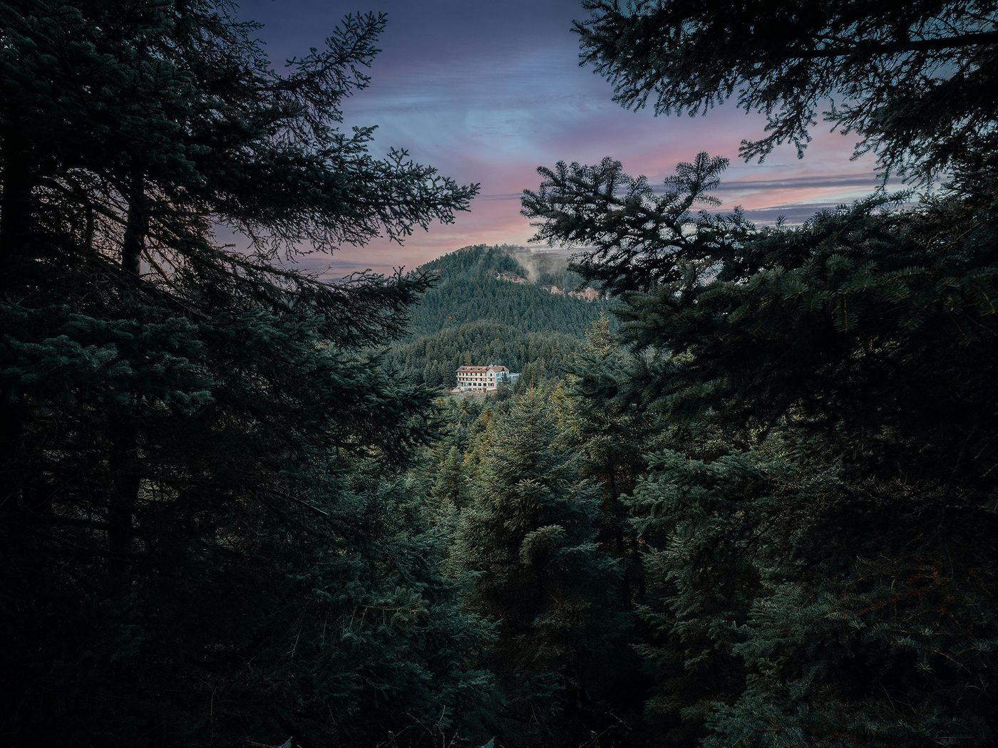 Framed by dark pine branches in the foreground, a classical hotel building with a red roof is nestled in the middle distance, surrounded by a sprawling forest under a dramatic pink and purple twilight sky. The scene conveys a sense of serene remoteness and timeless beauty. Framed by dark pine branches in the foreground, a classical hotel building with a red roof is nestled in the middle distance, surrounded by a sprawling forest under a dramatic pink and purple twilight sky. The scene conveys a sense of serene remoteness and timeless beauty.