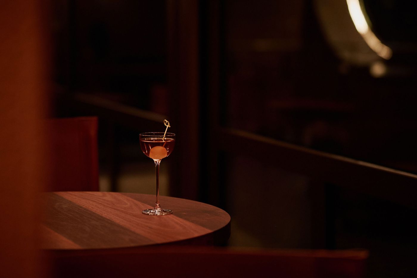 A stylized cocktail glass filled with a dark drink, garnished with a cherry, sits on a wooden table in a softly lit setting. A stylized cocktail glass filled with a dark drink, garnished with a cherry, sits on a wooden table in a softly lit setting.