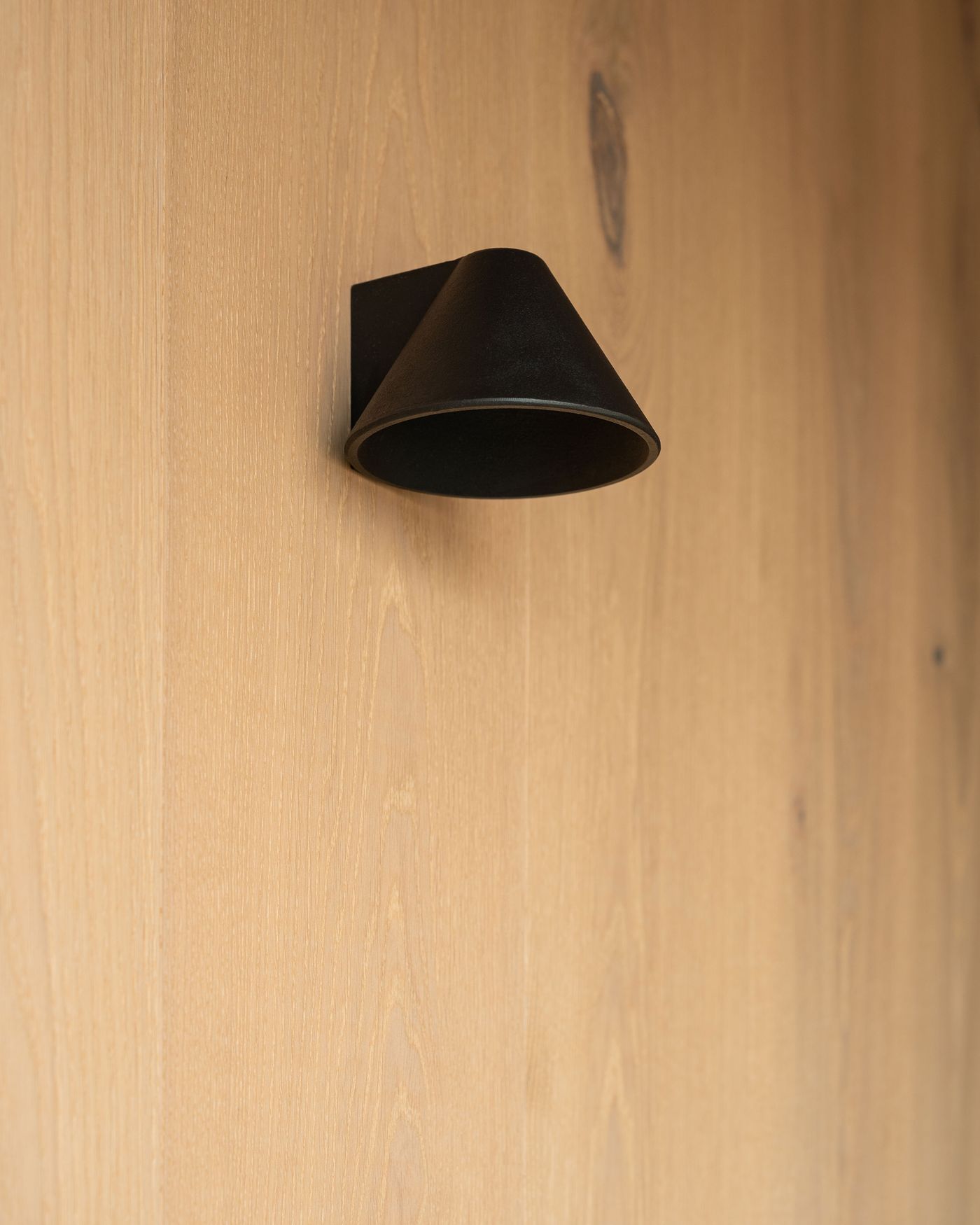 A black conical wall lamp mounted on a light wooden panel, showcasing minimalistic design and modern aesthetics.