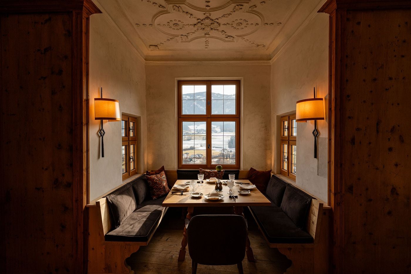 An intimate dining alcove at Castel Badia, framed like a stage by panelled jambs, sits beneath a delicate stucco ceiling of scrolls and garlands. A pine table set with cut-crystal glasses is wrapped by a charcoal velvet banquette, while flanking sconces and three timber windows look out over the village and snowy valley. An intimate dining alcove at Castel Badia, framed like a stage by panelled jambs, sits beneath a delicate stucco ceiling of scrolls and garlands. A pine table set with cut-crystal glasses is wrapped by a charcoal velvet banquette, while flanking sconces and three timber windows look out over the village and snowy valley.