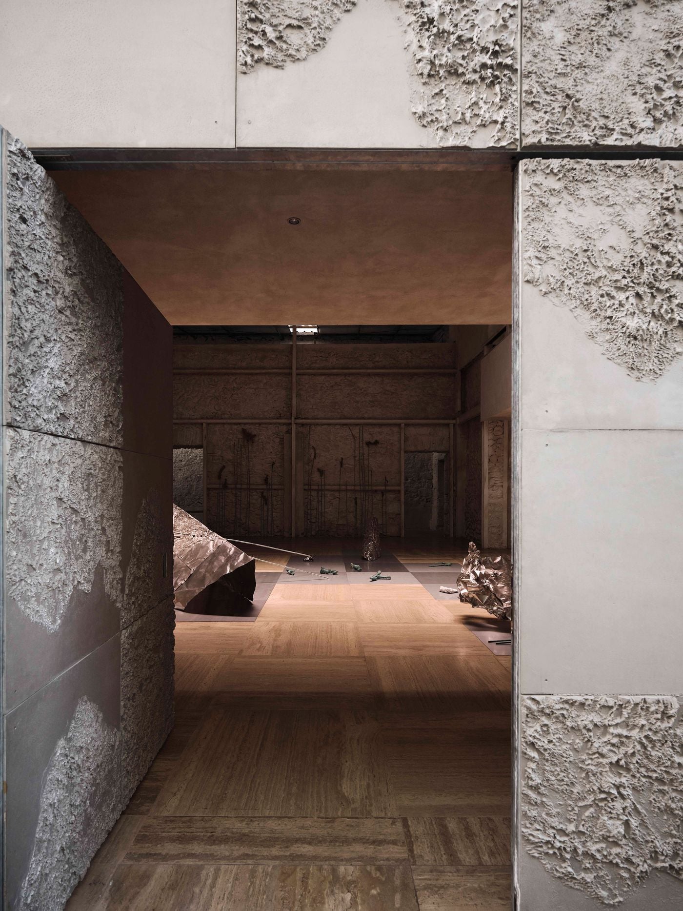 A doorway framed by metal panels with irregular, crust-like reliefs offers a layered view into the main hall and the “Casting Water” exhibition beyond. The composition encapsulates Castor Place’s material approach, setting rough masonry and tactile contemporary surfaces against the precise geometry of its architectural interventions. A doorway framed by metal panels with irregular, crust-like reliefs offers a layered view into the main hall and the “Casting Water” exhibition beyond. The composition encapsulates Castor Place’s material approach, setting rough masonry and tactile contemporary surfaces against the precise geometry of its architectural interventions.