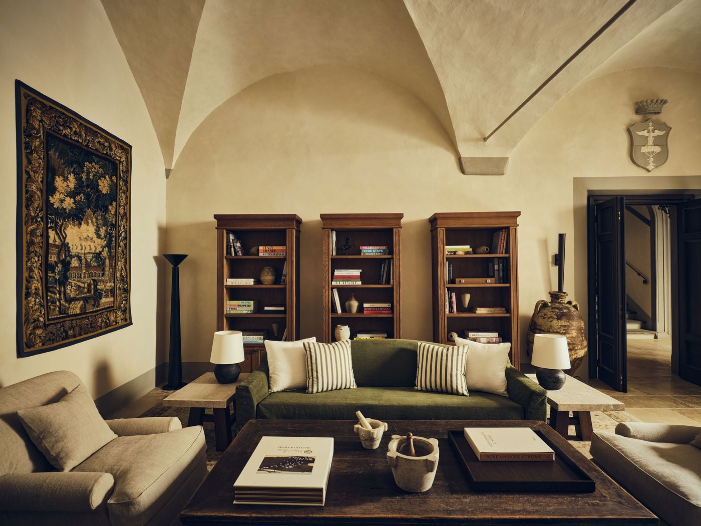 Three oak bookcases filled with travel and design titles line the vaulted lounge at La Réserve Firenze, behind a green linen sofa with striped cushions. A tall verdure tapestry, a weathered terracotta amphora, a second painted crest and marble mortars on a rustic coffee table layer the room with history and texture. Three oak bookcases filled with travel and design titles line the vaulted lounge at La Réserve Firenze, behind a green linen sofa with striped cushions. A tall verdure tapestry, a weathered terracotta amphora, a second painted crest and marble mortars on a rustic coffee table layer the room with history and texture.