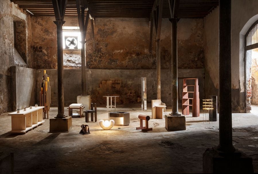 The "Perspective and Matter" exhibition by Luso Collective transforms a raw, industrial space into a gallery for contemporary furniture. An array of minimalist designs in various materials like wood, metal, and stone are artfully arranged amidst the decaying concrete walls and exposed timber beams, creating a dramatic interplay between raw architecture and refined objects.