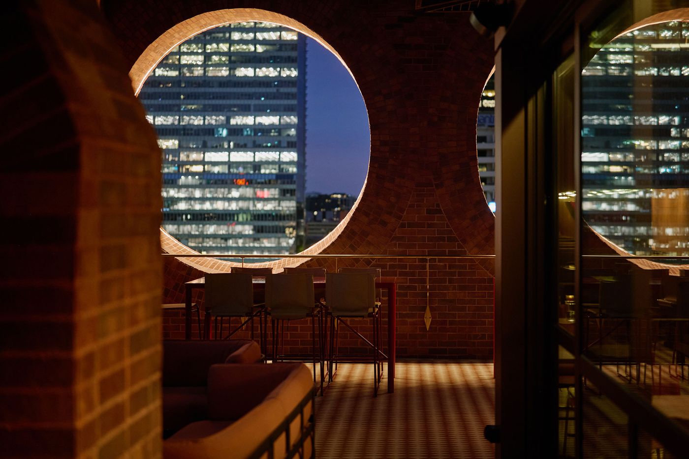 A cozy terrace with circular windows overlooking a city skyline at night, featuring a seating area and modern bar stools. A cozy terrace with circular windows overlooking a city skyline at night, featuring a seating area and modern bar stools.