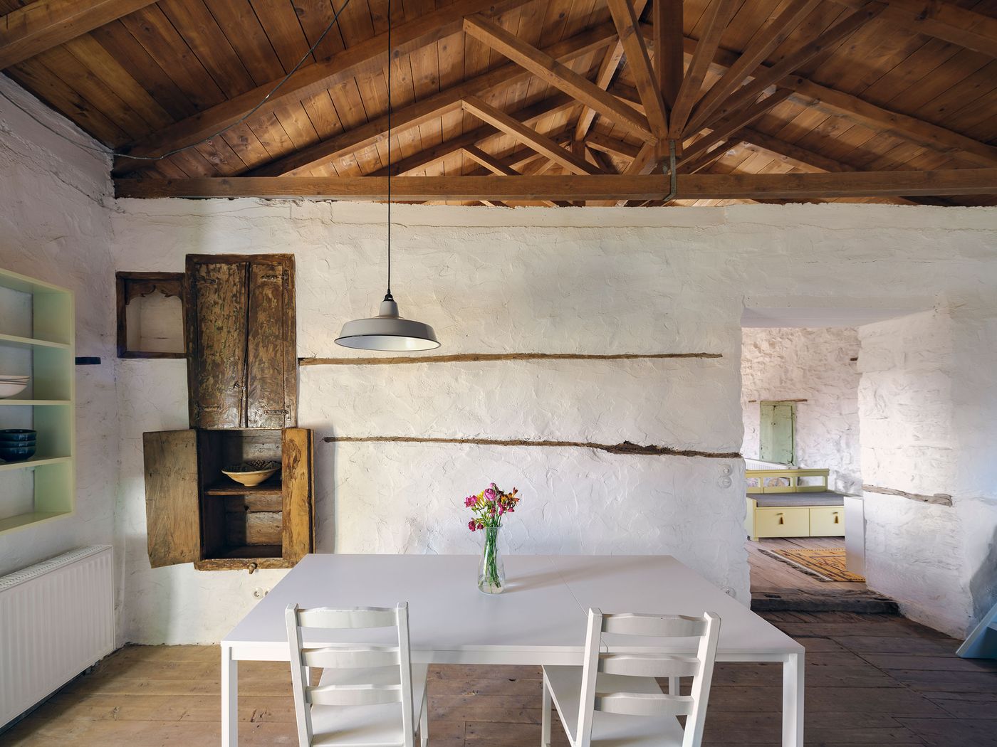 Traditional wooden cabinet creates authentic storage within the whitewashed dining area, where exposed timber ceiling and simple white furniture celebrate the building's rustic Mediterranean heritage. The composition demonstrates how contemporary restoration can preserve original character while providing necessary modern amenities through careful material selection.