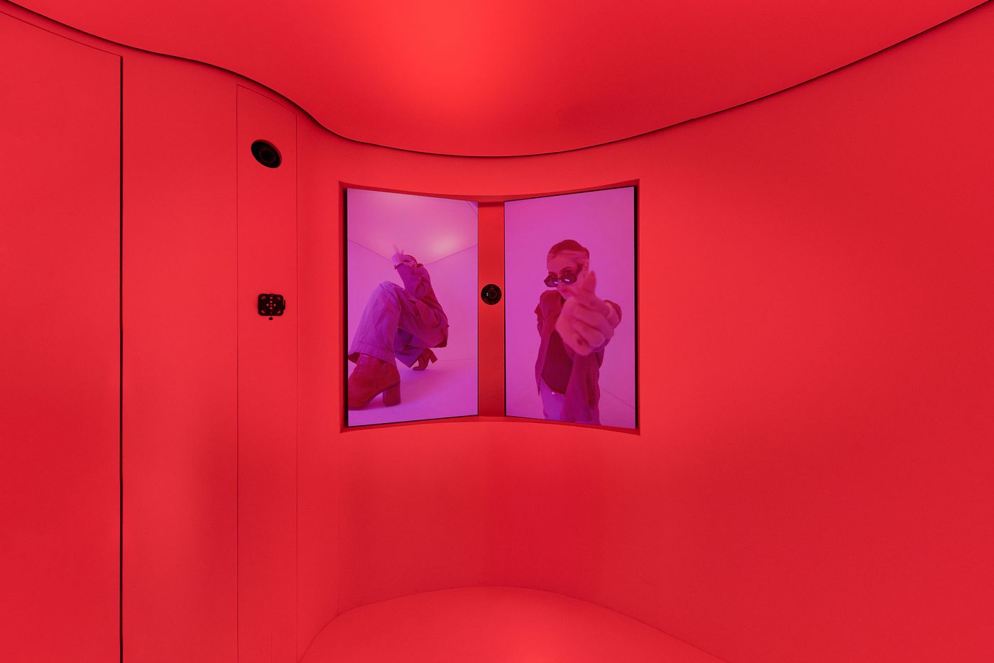 A vibrant red room features two display screens showing individuals in striking outfits and poses, creating a dynamic visual experience. A vibrant red room features two display screens showing individuals in striking outfits and poses, creating a dynamic visual experience.