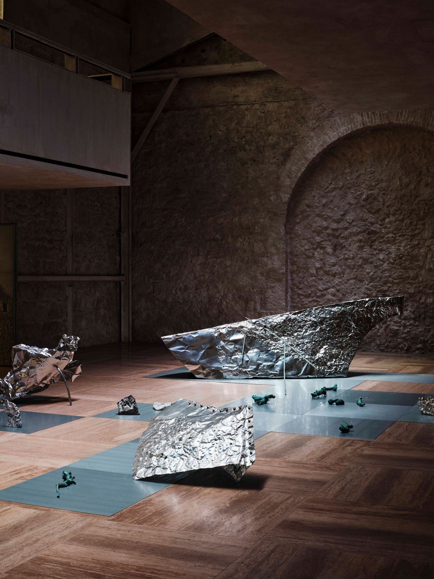 Reflective sculptural forms from “Casting Water” occupy Castor Place’s travertine floor, their crumpled metallic surfaces catching focused light against the subdued masonry backdrop. Grey floor panels weave between the works, while a large bricked-up arch and suspended gallery preserve a legible record of the building’s previous configurations. Reflective sculptural forms from “Casting Water” occupy Castor Place’s travertine floor, their crumpled metallic surfaces catching focused light against the subdued masonry backdrop. Grey floor panels weave between the works, while a large bricked-up arch and suspended gallery preserve a legible record of the building’s previous configurations.