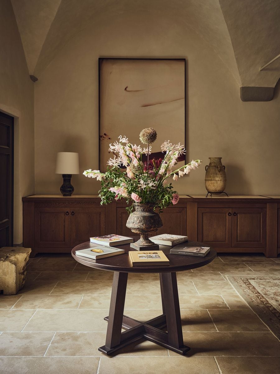 A round dark oak table at La Réserve Firenze holds monographs on Brancusi and Donatello around an antique urn spilling with pale pink blooms. Behind it, a large abstract canvas of a single sweeping gesture hangs above a long oak sideboard, with an amphora on an iron stand and a rough-hewn stone fragment nearby. A round dark oak table at La Réserve Firenze holds monographs on Brancusi and Donatello around an antique urn spilling with pale pink blooms. Behind it, a large abstract canvas of a single sweeping gesture hangs above a long oak sideboard, with an amphora on an iron stand and a rough-hewn stone fragment nearby.