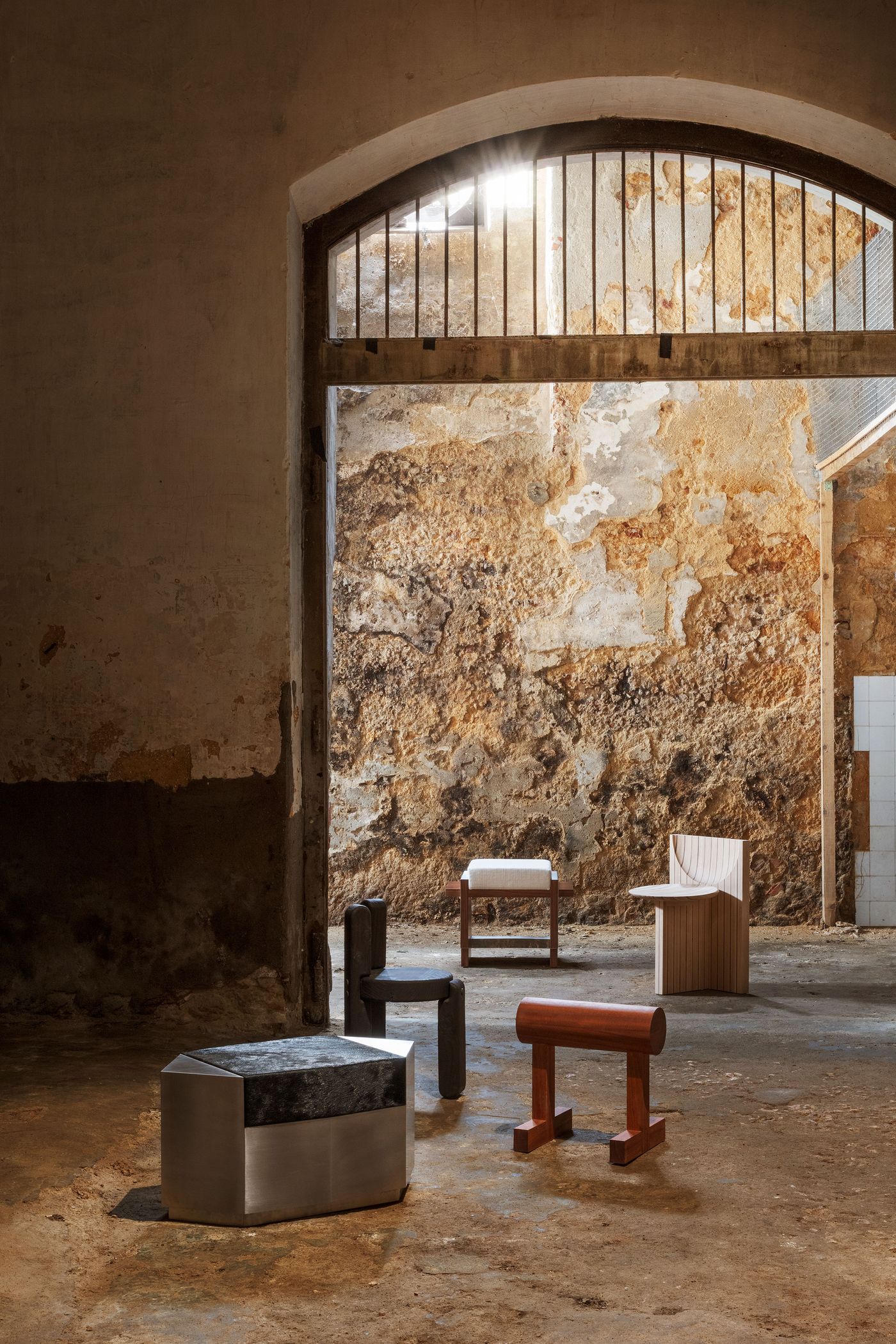 An evocative scene from the "Perspective and Matter" exhibition by Luso Collective, set within a grand, raw industrial archway. Various contemporary furniture pieces, including a metal and stone bench, a wooden stool, and a sculptural chair, are strategically placed amidst the decaying concrete and stone walls, highlighting a brutalist influence and the beauty of natural materials. An evocative scene from the "Perspective and Matter" exhibition by Luso Collective, set within a grand, raw industrial archway. Various contemporary furniture pieces, including a metal and stone bench, a wooden stool, and a sculptural chair, are strategically placed amidst the decaying concrete and stone walls, highlighting a brutalist influence and the beauty of natural materials.