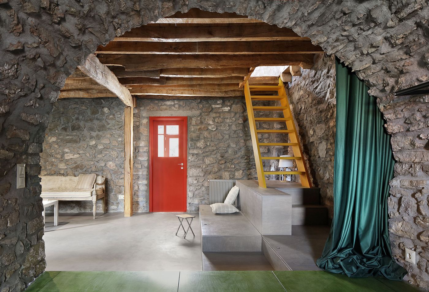 Dramatic stone archway frames the living area where coral-red door provides vibrant contemporary accent against the raw limestone walls. The yellow staircase creates bold architectural statement while emerald-green drapery adds theatrical warmth, demonstrating how color can activate traditional spaces without compromising their authentic material character.