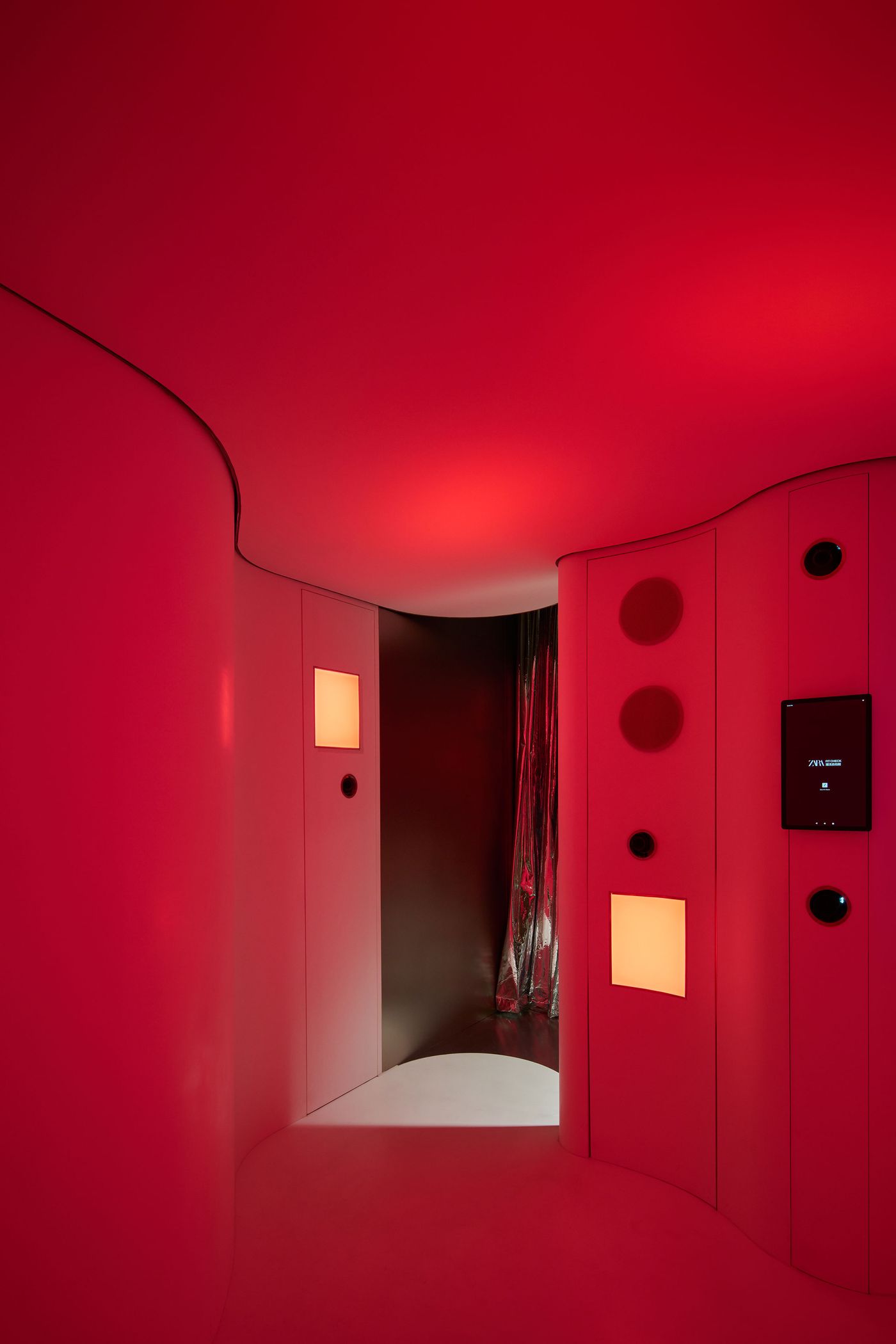 A sleek, modern hallway illuminated in red, featuring curved walls, speakers, and a touchscreen panel, leading to a darkened space. A sleek, modern hallway illuminated in red, featuring curved walls, speakers, and a touchscreen panel, leading to a darkened space.
