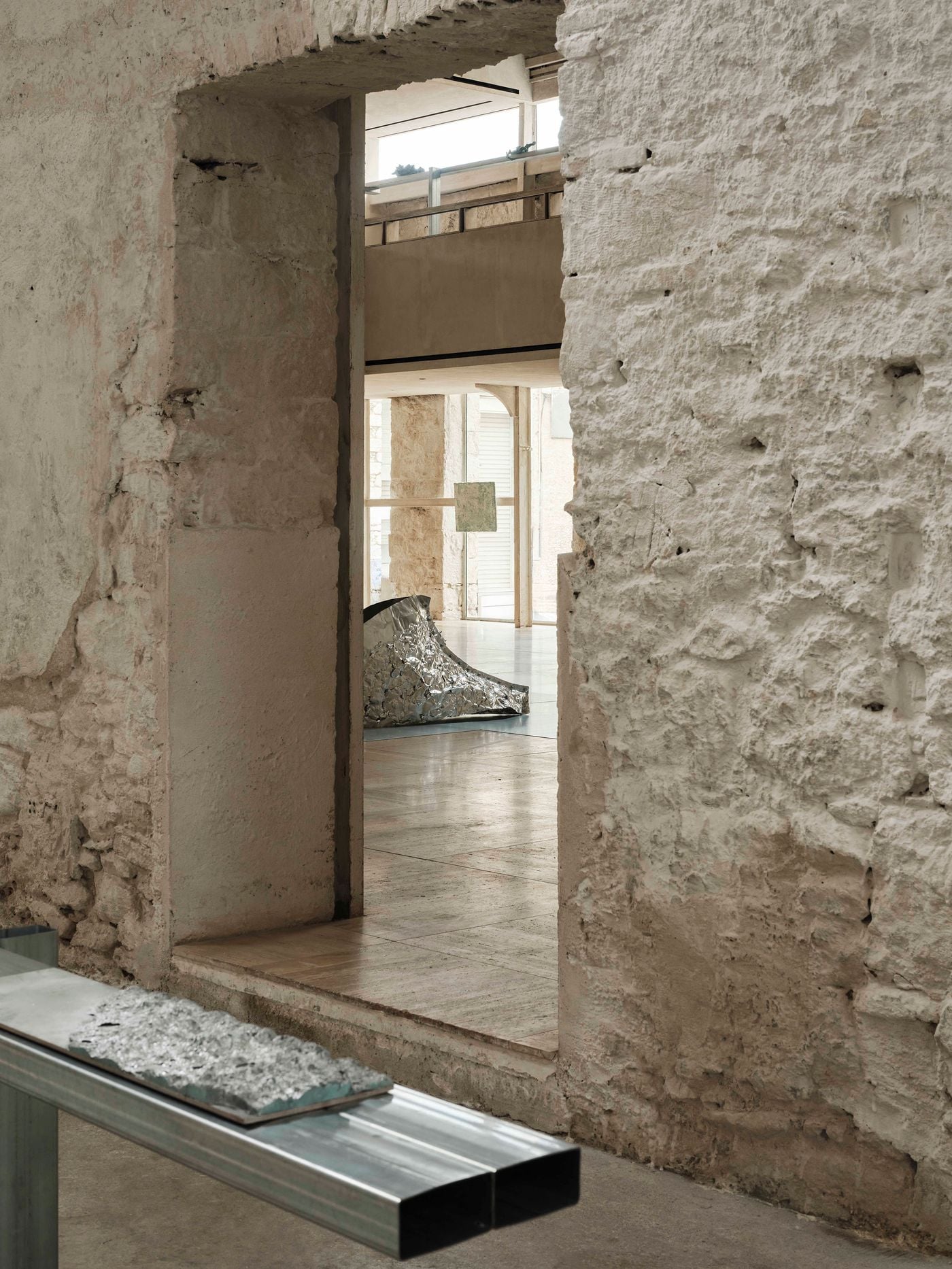 A rough-cut doorway creates a tightly framed view from one of Castor Place’s smaller rooms into the brighter main hall, where a metallic work from “Casting Water” is partially visible. Exposed stone, patched plaster and a minimal steel display table foreground the project’s dialogue between retained fabric and contemporary intervention. A rough-cut doorway creates a tightly framed view from one of Castor Place’s smaller rooms into the brighter main hall, where a metallic work from “Casting Water” is partially visible. Exposed stone, patched plaster and a minimal steel display table foreground the project’s dialogue between retained fabric and contemporary intervention.