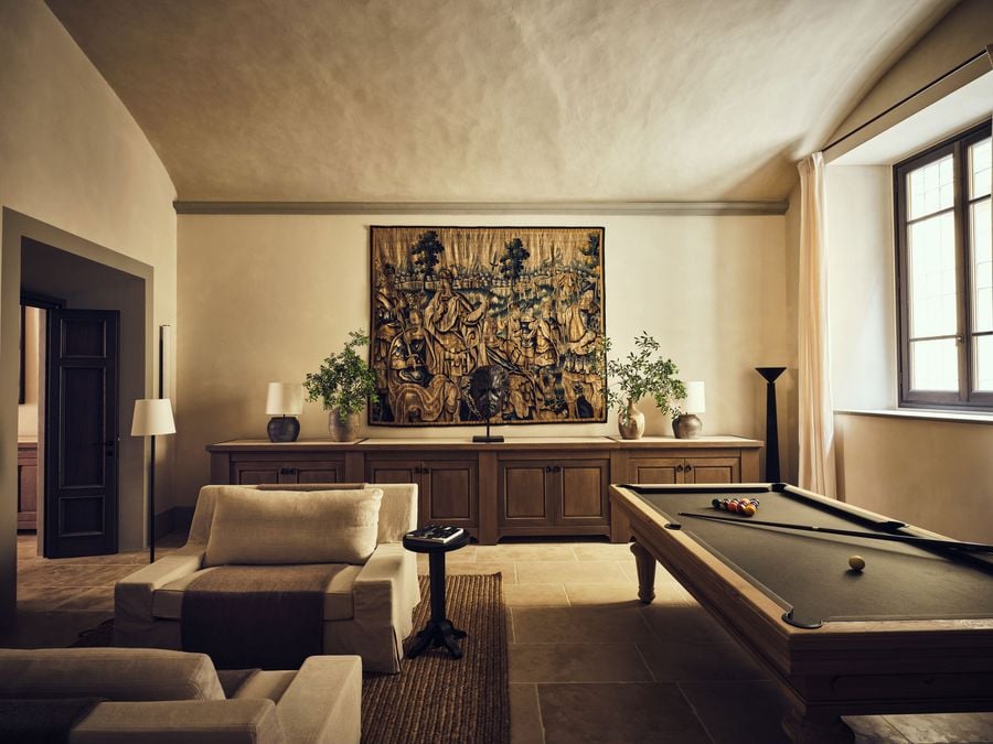 The games room at La Réserve Firenze sets an oak billiard table beneath a gently vaulted ceiling and an antique tapestry of armoured figures. A dark bronze head on a stand, a long oak sideboard with earthenware lamps, linen armchairs and a slender torchère frame a space for unhurried evenings.
