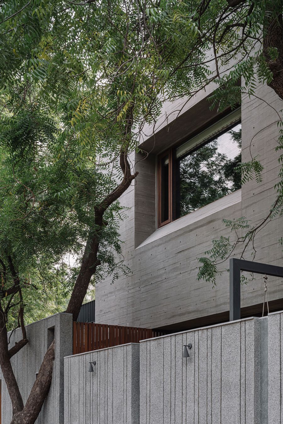 An angular view of MS House's concrete exterior, softened by the dense canopy of trees. The raw concrete walls feature subtle textures, and a large window reflects the surrounding foliage. A minimalist concrete wall with vertical grooves and subtle outdoor lighting fixtures frames the foreground, showcasing a blend of brutalist design and natural integration.