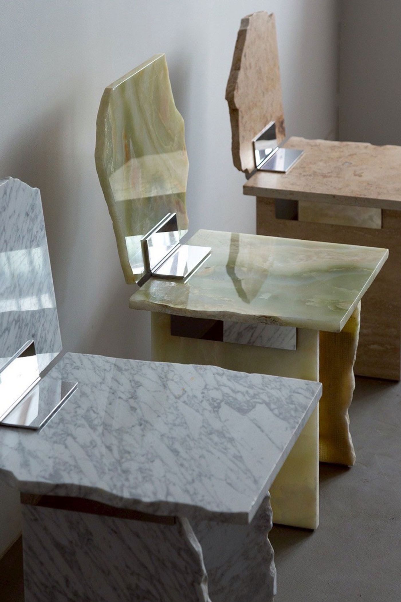 Three sculptural chairs made from marble and onyx with angular backs, featuring sleek metal brackets against a neutral wall. Three sculptural chairs made from marble and onyx with angular backs, featuring sleek metal brackets against a neutral wall.