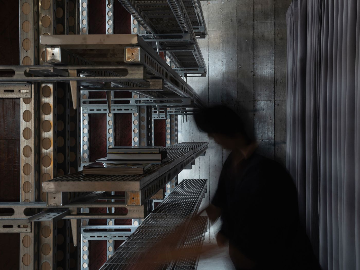 An intriguing detail shot from 889 GLO Art Space showcases a multi-tiered shelving system crafted from industrial metal components, displaying books and objects. The raw, brutalist concrete wall and dark, heavy curtains provide a stark backdrop, emphasizing the intricate, modular design and the space's industrial aesthetic. A person in motion adds a sense of human interaction.