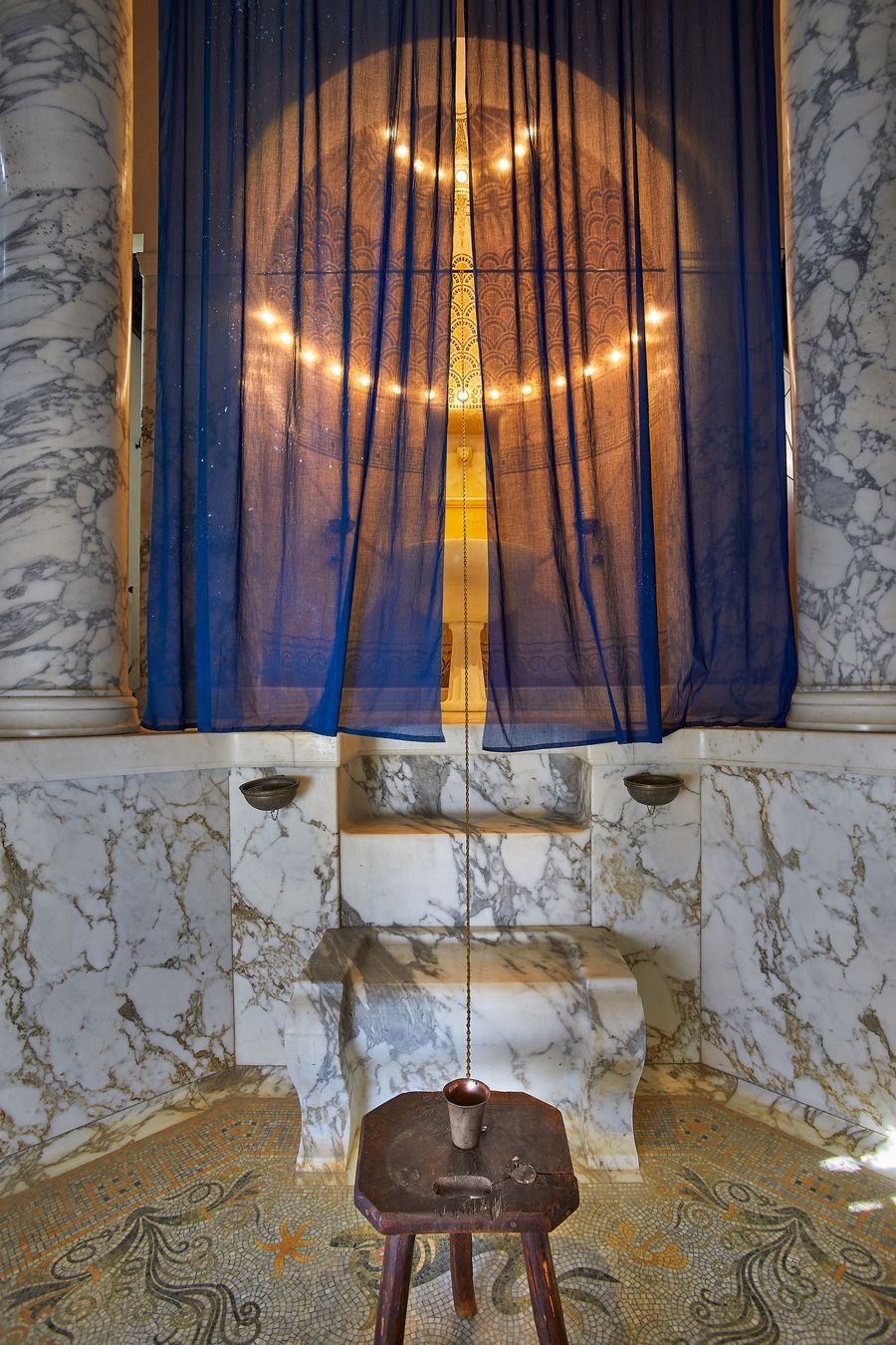 A classical bathing space at Villa Kerylos with large marble columns and an ornate mosaic floor. A dark blue translucent curtain hangs in front of a circular, illuminated art piece. A small, rustic wooden stool sits centrally, holding a copper vessel, creating a stark contrast against the refined architectural details. A classical bathing space at Villa Kerylos with large marble columns and an ornate mosaic floor. A dark blue translucent curtain hangs in front of a circular, illuminated art piece. A small, rustic wooden stool sits centrally, holding a copper vessel, creating a stark contrast against the refined architectural details.