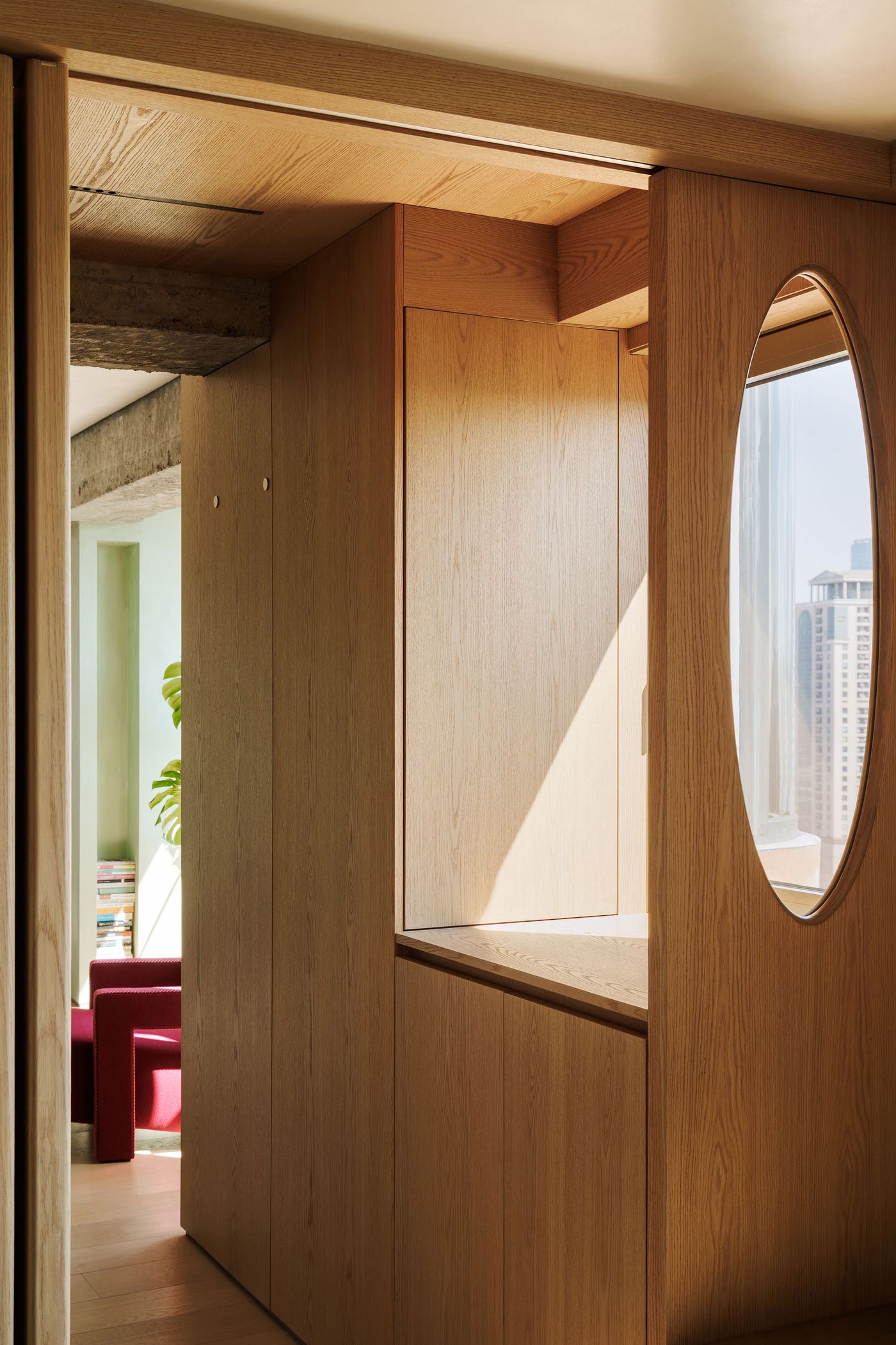 An elegant hallway clad in light wood paneling, featuring a distinctive oval cut-out window that frames a glimpse of the cityscape. The play of natural light creates dynamic shadows, emphasizing the clean lines and meticulous craftsmanship of this contemporary design. Beyond, a vibrant red armchair hints at a more expansive living area.