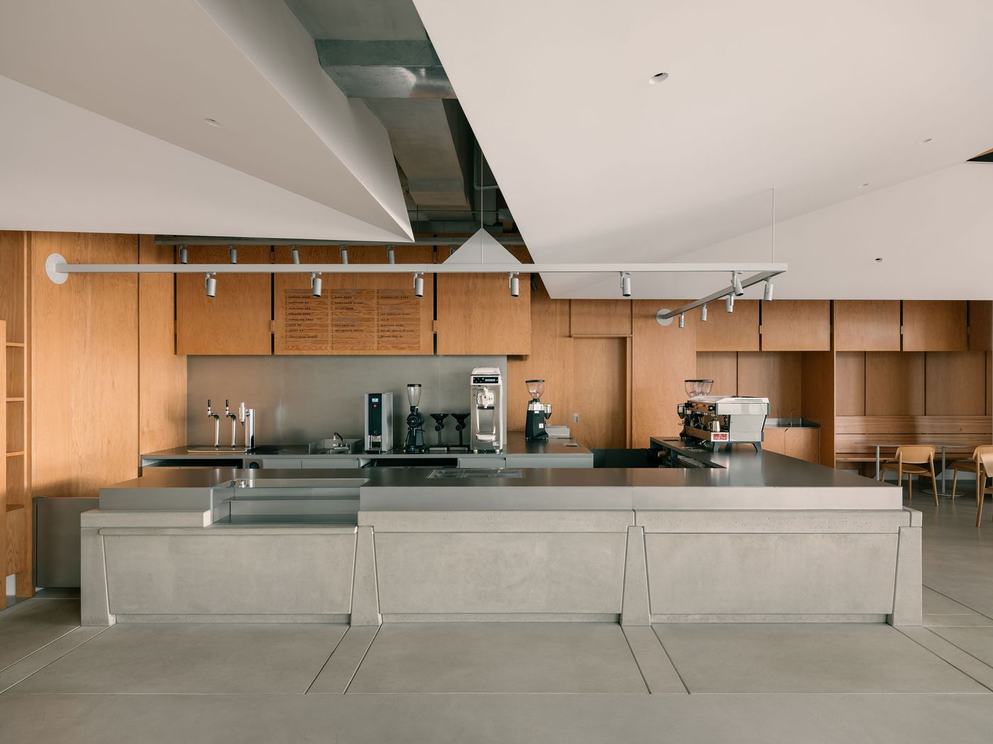 The barista counter takes center stage, rendered in angular concrete volumes and backed by warm wooden panels and exposed ductwork, blending industrial edge with architectural refinement.